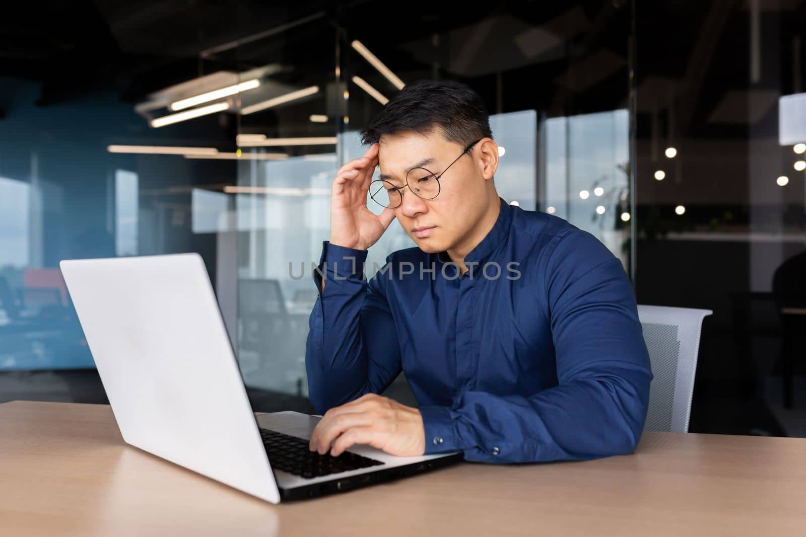 Thoughtful and serious male Asian student sits in the office at the table, works on a laptop. Passes the exam, worries, studies online through a video call. He holds his head with his hand.