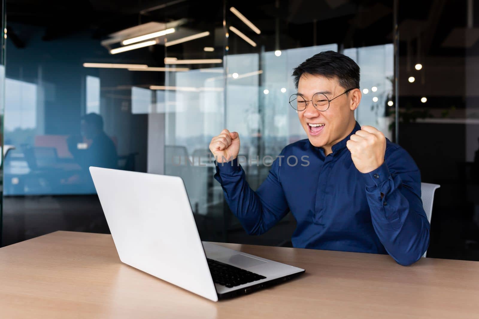 A happy young Asian student man sits in the office at the table, looks at the laptop and rejoices. Passed the exam, received a letter, accepted a job, entered the university. Screams with joy.