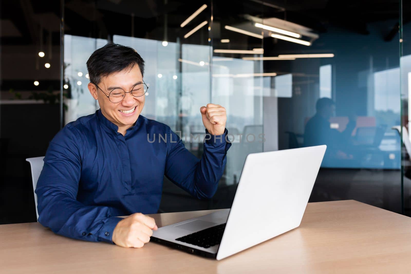 A young Asian man is working in the office on a laptop online. He is happy, looks at the monitor, makes a yes gesture with his hand.