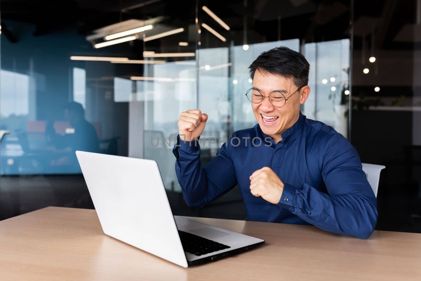 A young male Asian student received good news, mail, passed the exam, entered the university, received a scholarship. He sits in the office, looks at the monitor, rejoices, shows yes with his hand