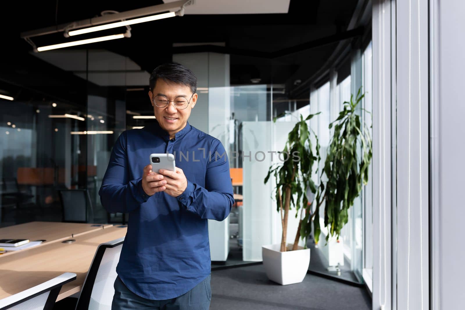A young man, an Asian businessman, a student standing by the window in the office, uses a mobile phone, types messages, reads, checks mail. He looks at the camera, smiles.