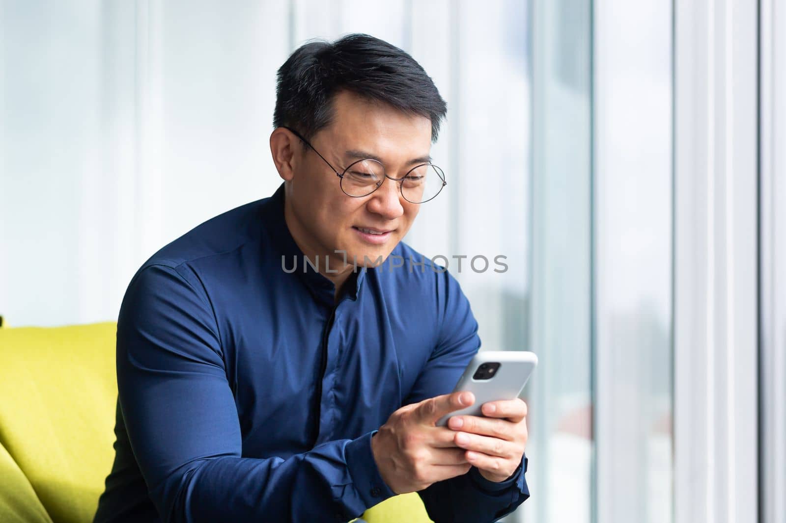 A young man, an Asian student, sits on an ottoman by the window in an office, co-working space, campus. He holds the phone in his hands, dials a message, reads the news, checks the mail, smiles.