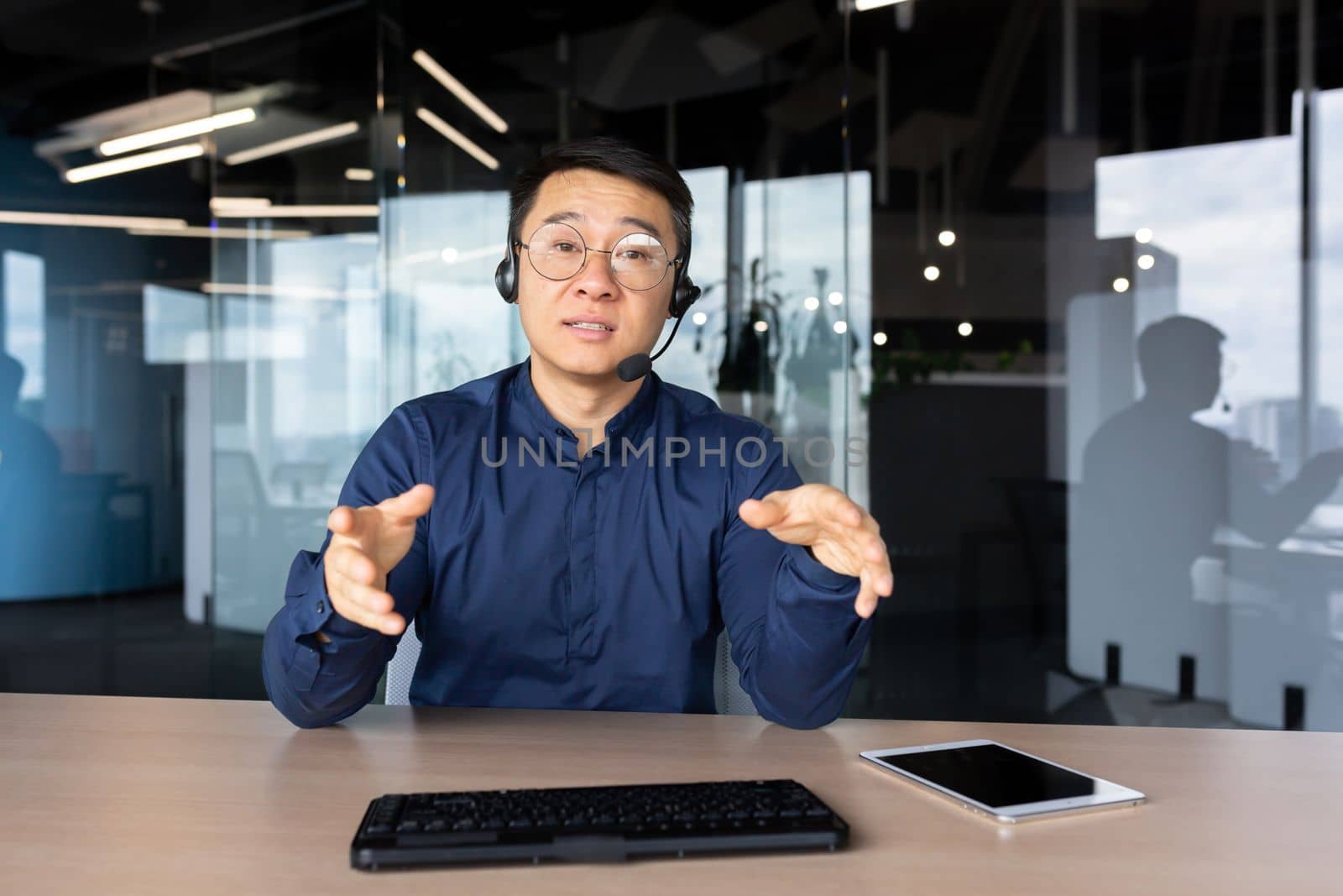 A young Asian man sits in the office at a desk in a headset, looks and talks to the camera. Explains, advises, conducts training, online meeting.