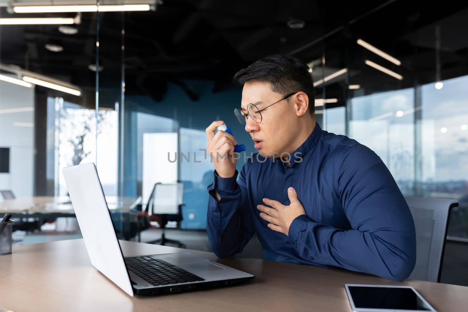 Young male Asian office worker having asthma attack at workplace, breathing hard. He sits at a table with a laptop, uses an inhaler.