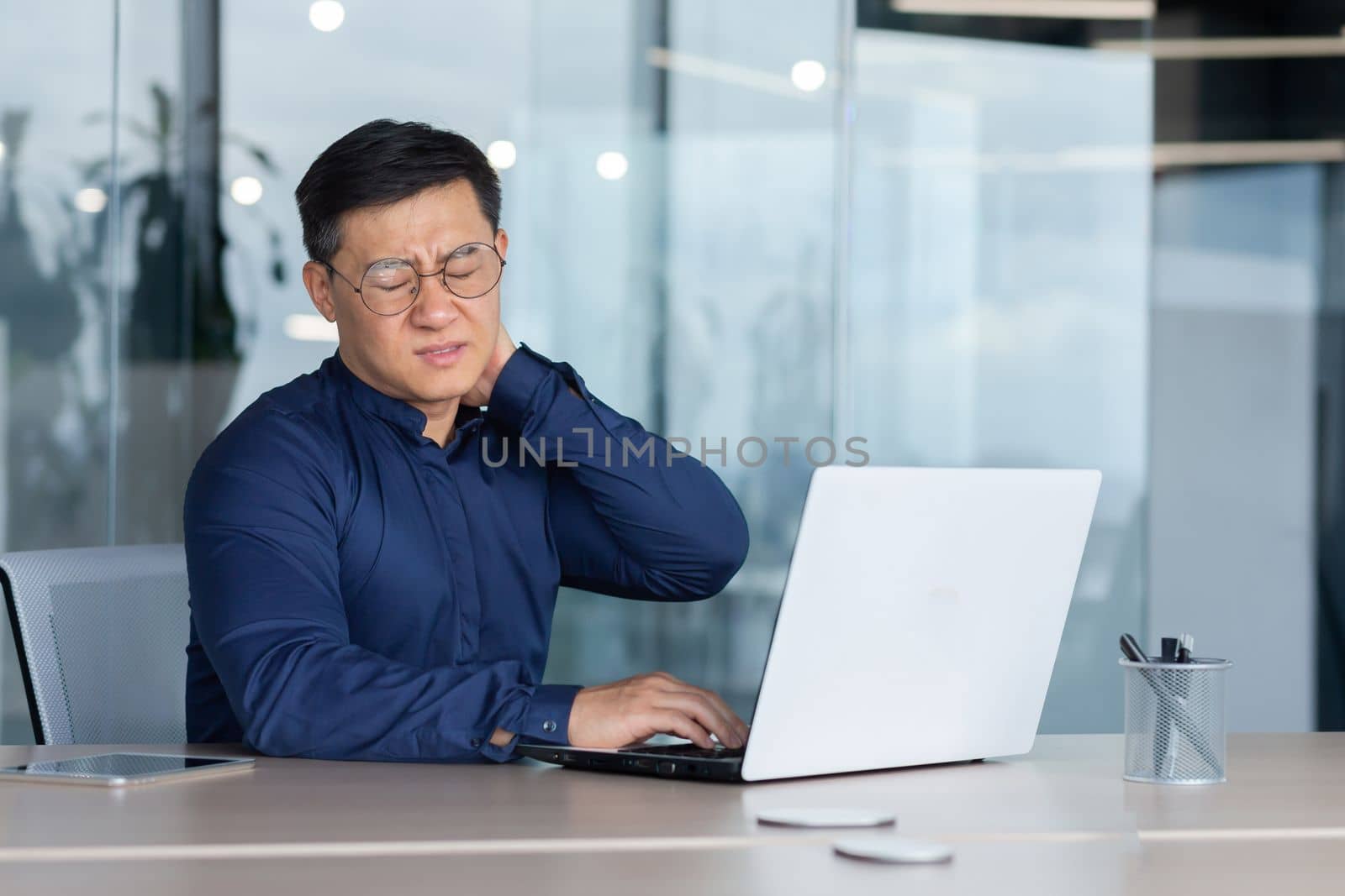 A tired young man, a freelancer, a businessman, sits behind a desk in the office, feels unbearable pain in his neck, closes his eyes, grimaces in pain, massages his hand.