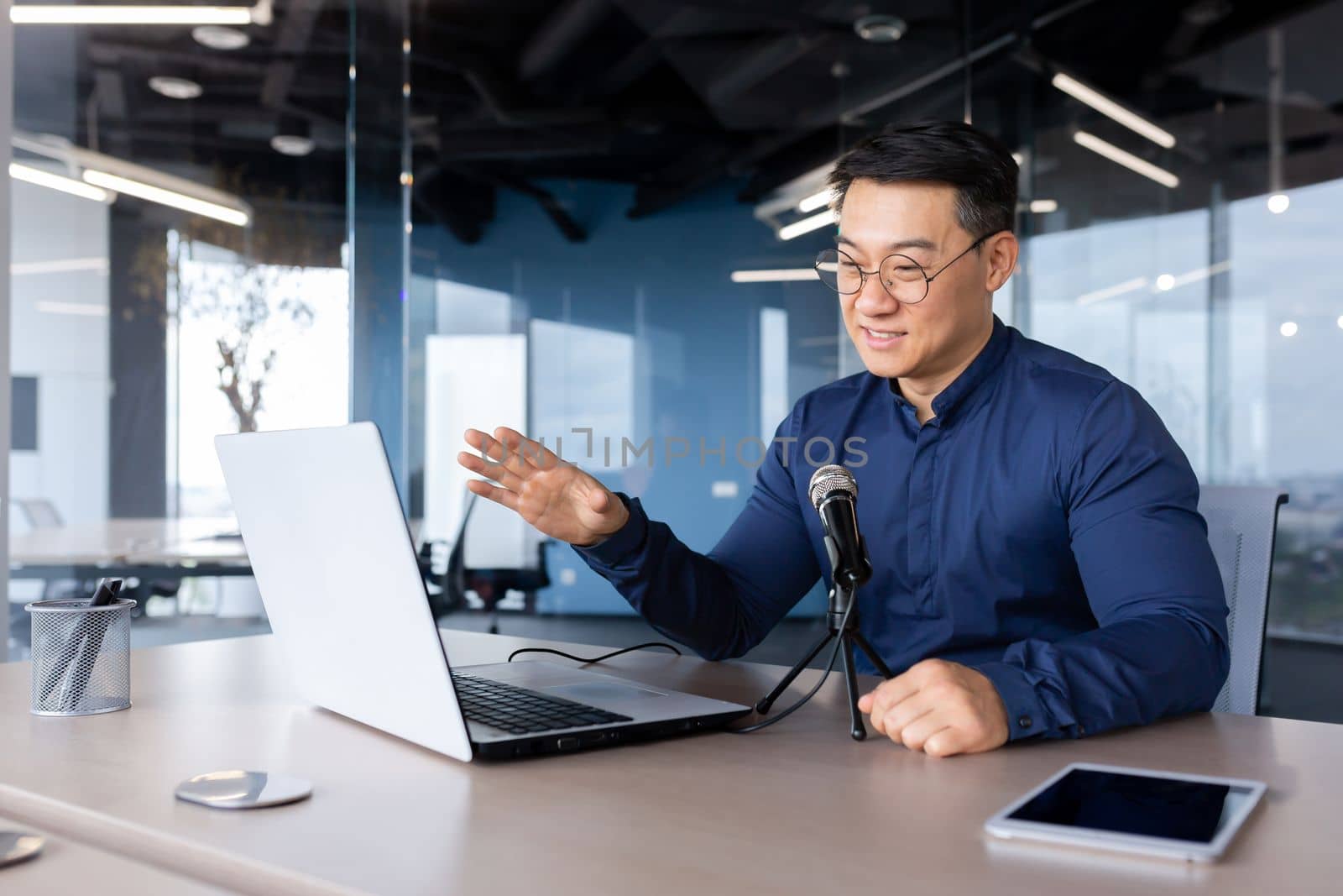 A young Asian man is sitting at a desk in the office. Speaks into the microphone through a video call from a laptop. Online business meeting, blogger, recording interviews, podcast.