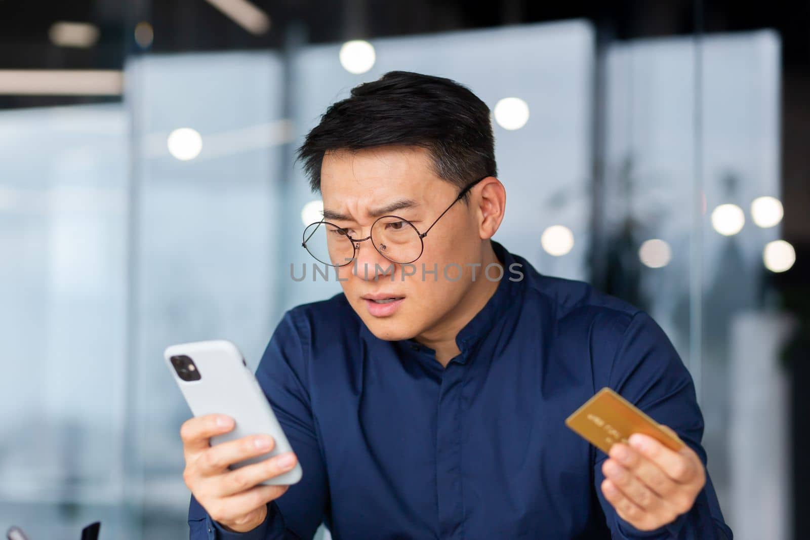Close-up photo. A worried young Asian man in glasses holds a credit card and look shocked at the phone. Checks account, failed online purchases, bankruptcy.