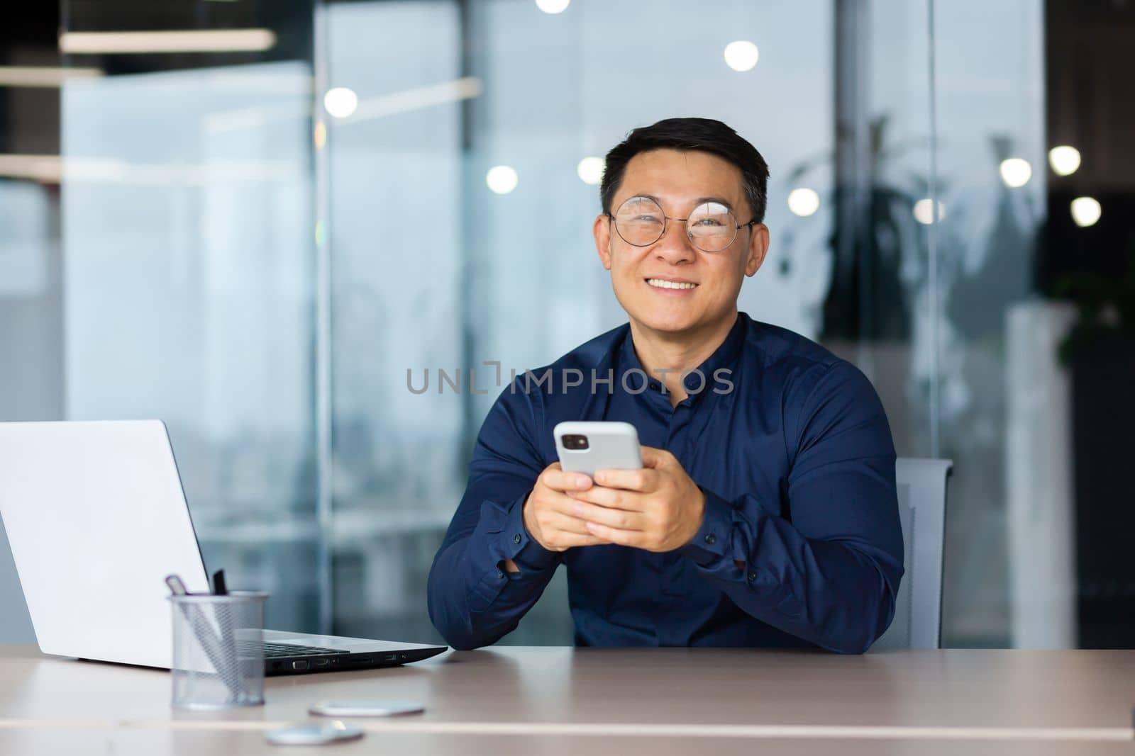 A young man in glasses sits in the office at a table with a laptop, holds a phone in his hands, smiles at the camera.