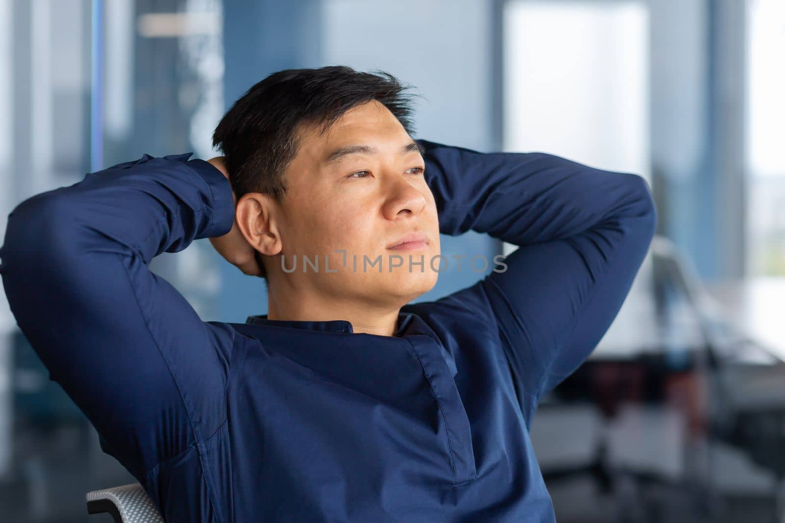 A calm young Asian man sits in the office on a chair, leans back, hands behind his head, looks thoughtfully and dreamily out the window. Resting from a hard day, satisfied with successful work.