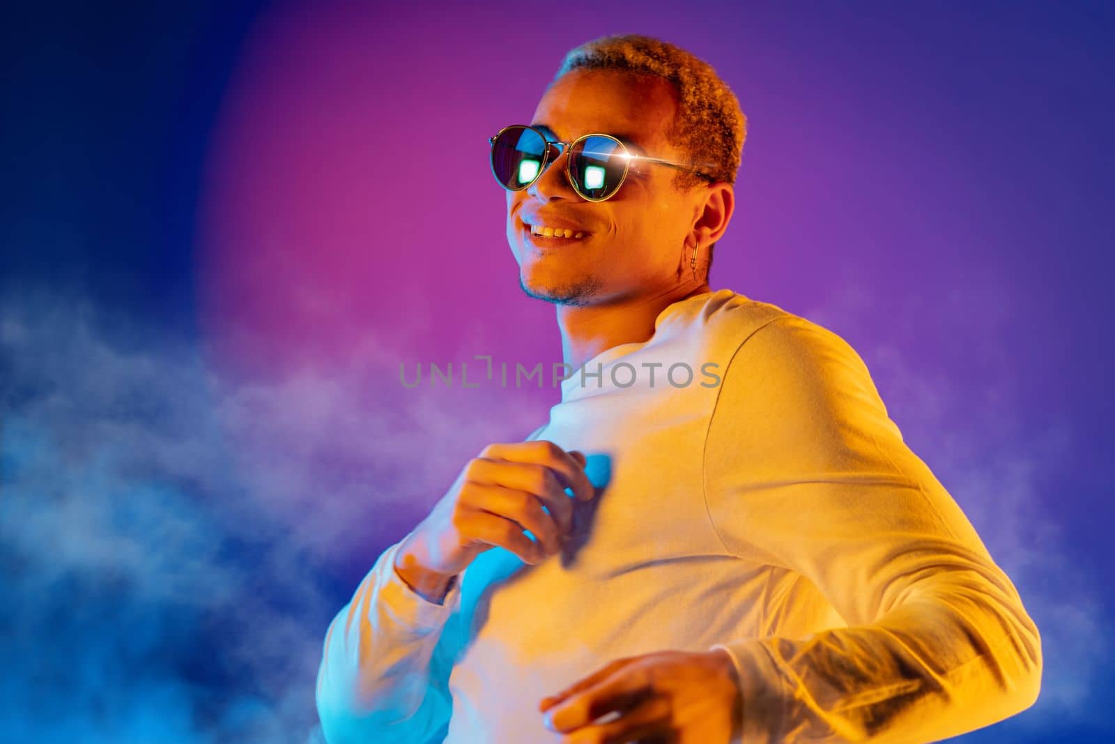 Energetic african american man dancing on colorful background under neon light. Portrait of stylish guy with dyed white curly hairstyle and round mirror eyewear. High quality photo