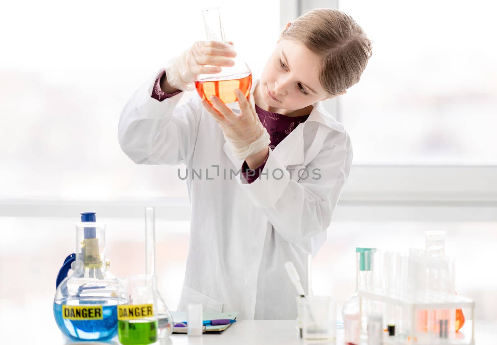 Smart girl during scientific chemistry experiment wearing protection glasses, holding bottle with orange liquid. Schoolgirl with chemical equipment on school lesson portrait