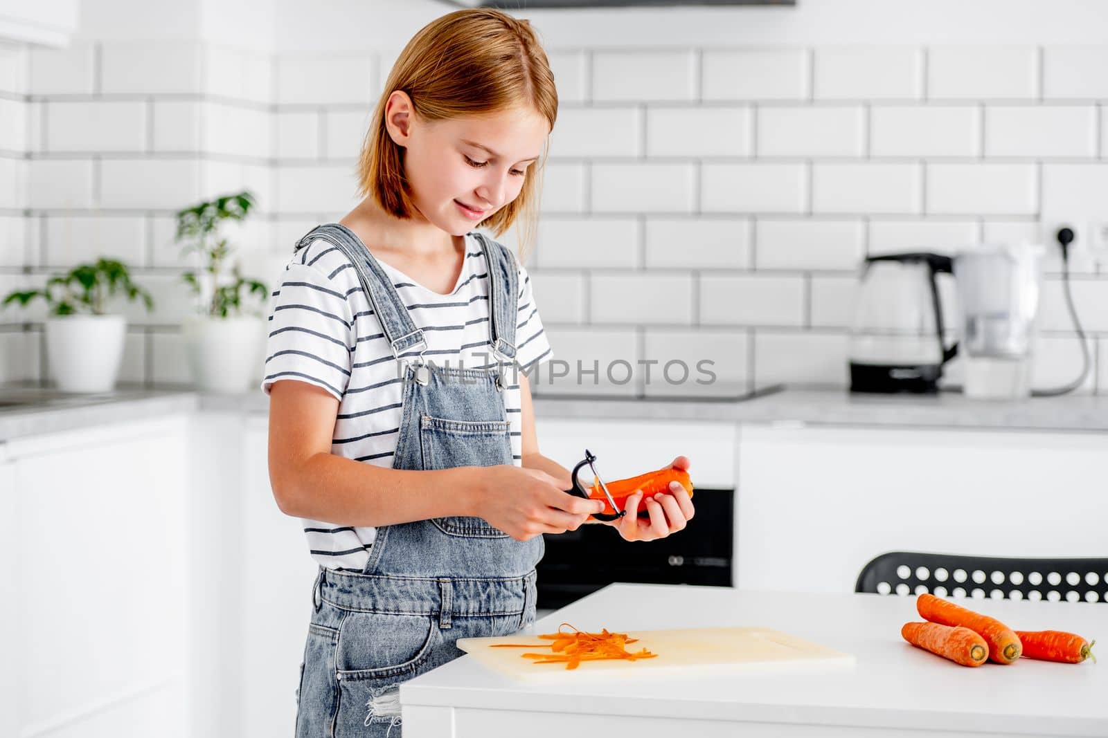 Preteen girl pilling carrot at kitchen and smiling. Child with orange vegetables food cooking at home