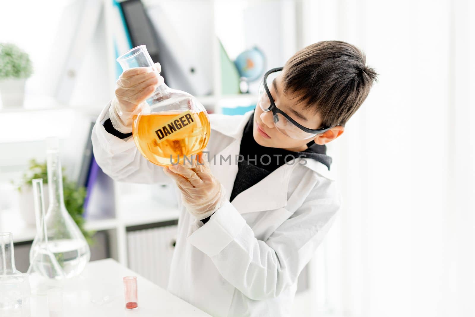 School boy wearing protection glasses doing chemistry experiment in elementary science class. Clever pupil holding tube with danger mark in lab during test