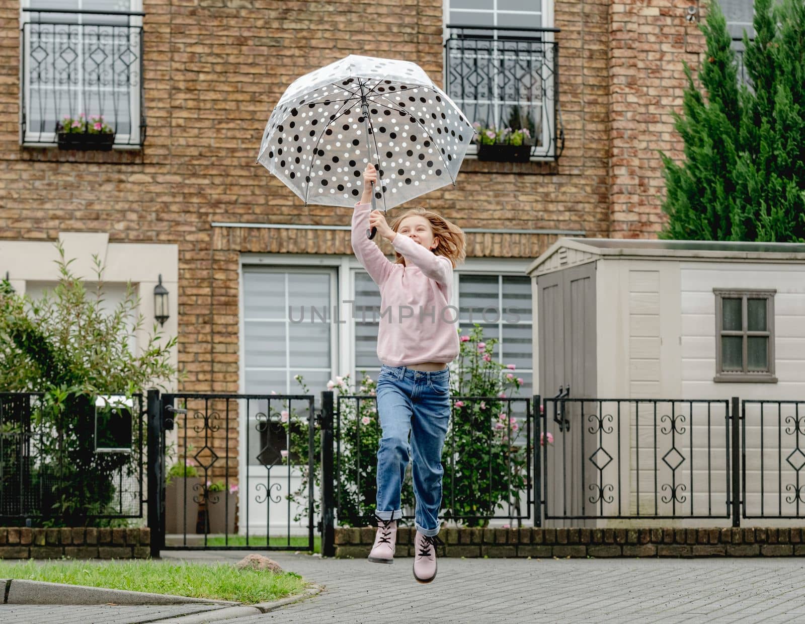 Preteen girl with umbrella running and jumping outdoors. Pretty child kid portrait in rainy day at street