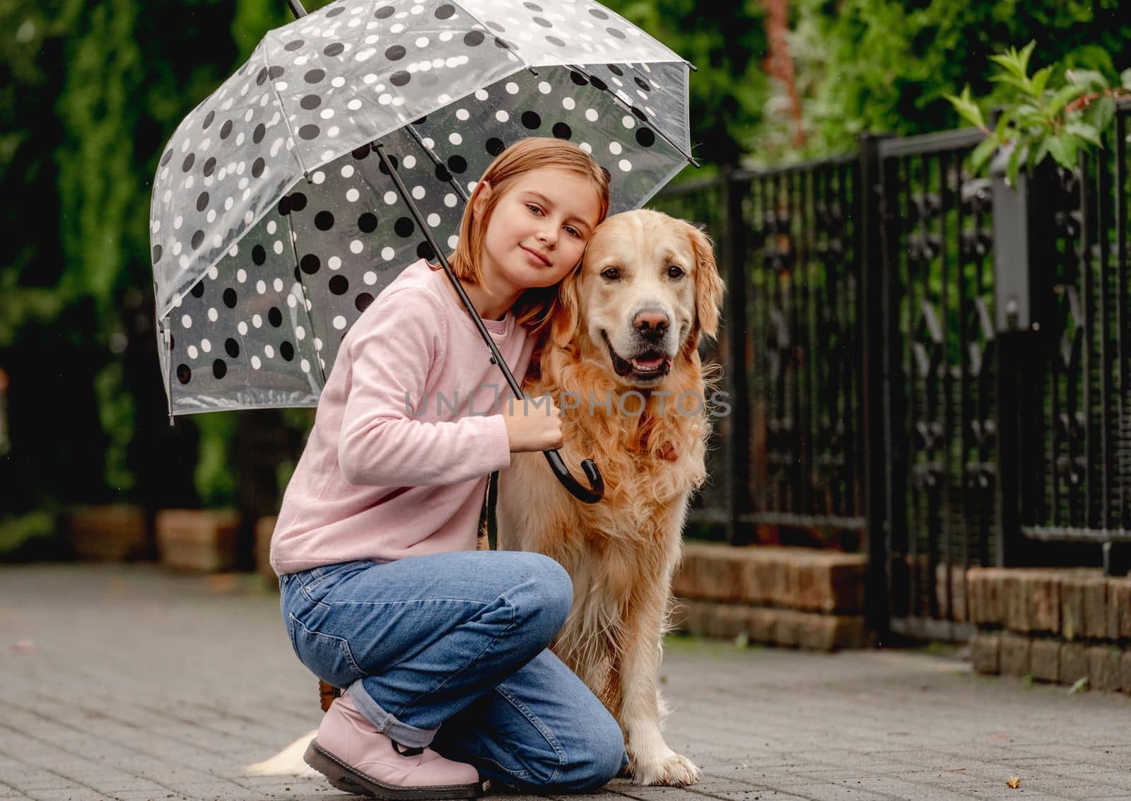 Preteen girl with golden retriever dog sitting under umbrella at street and looking back. Pretty kid child with doggy pet in rainy day