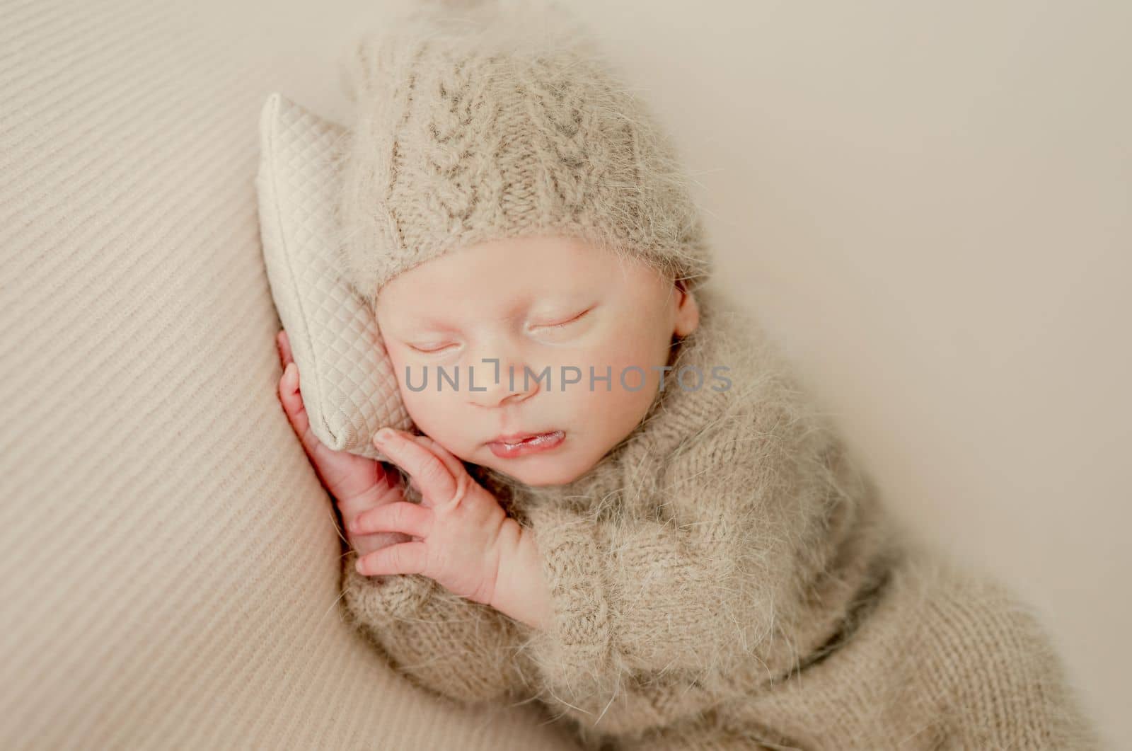 Newborn baby boy sleeping and holding hands under cheeks. Adorable infant child kid napping wearing knitted beige hat and costume