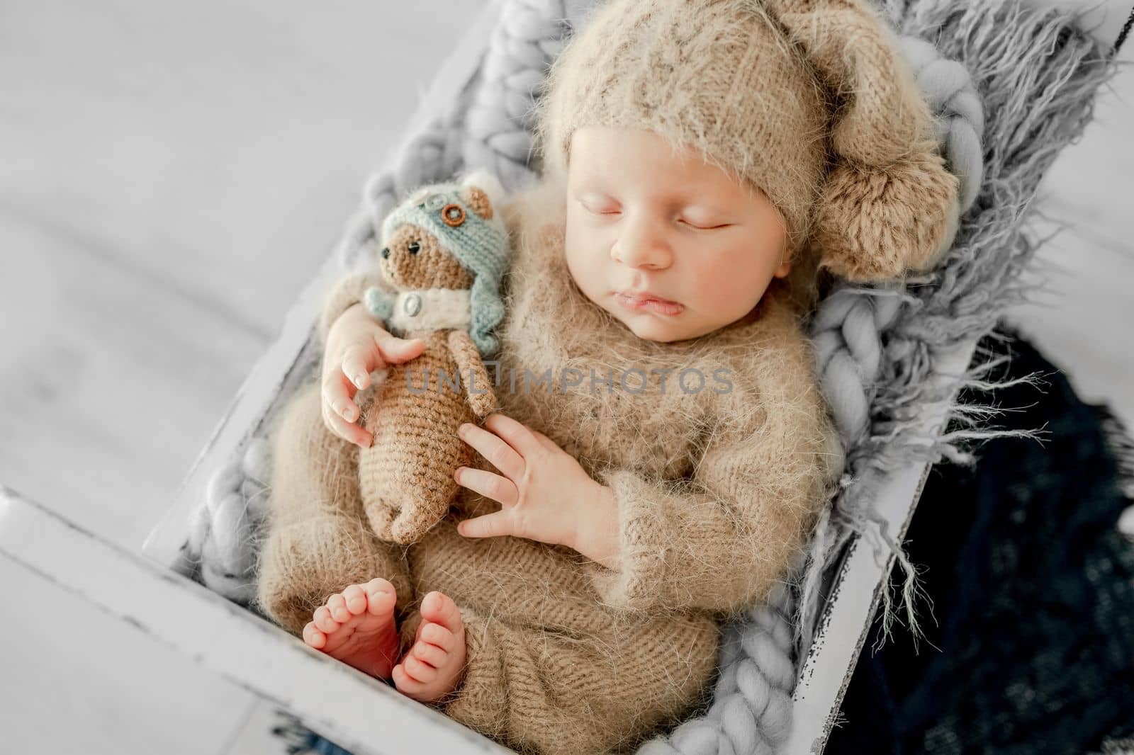 Newborn baby boy sleeping on fur in tiny bed and holding hands under cheeks. Adorable infant child kid napping on his tummy during studio photoshoot wearing knitted hat and costume