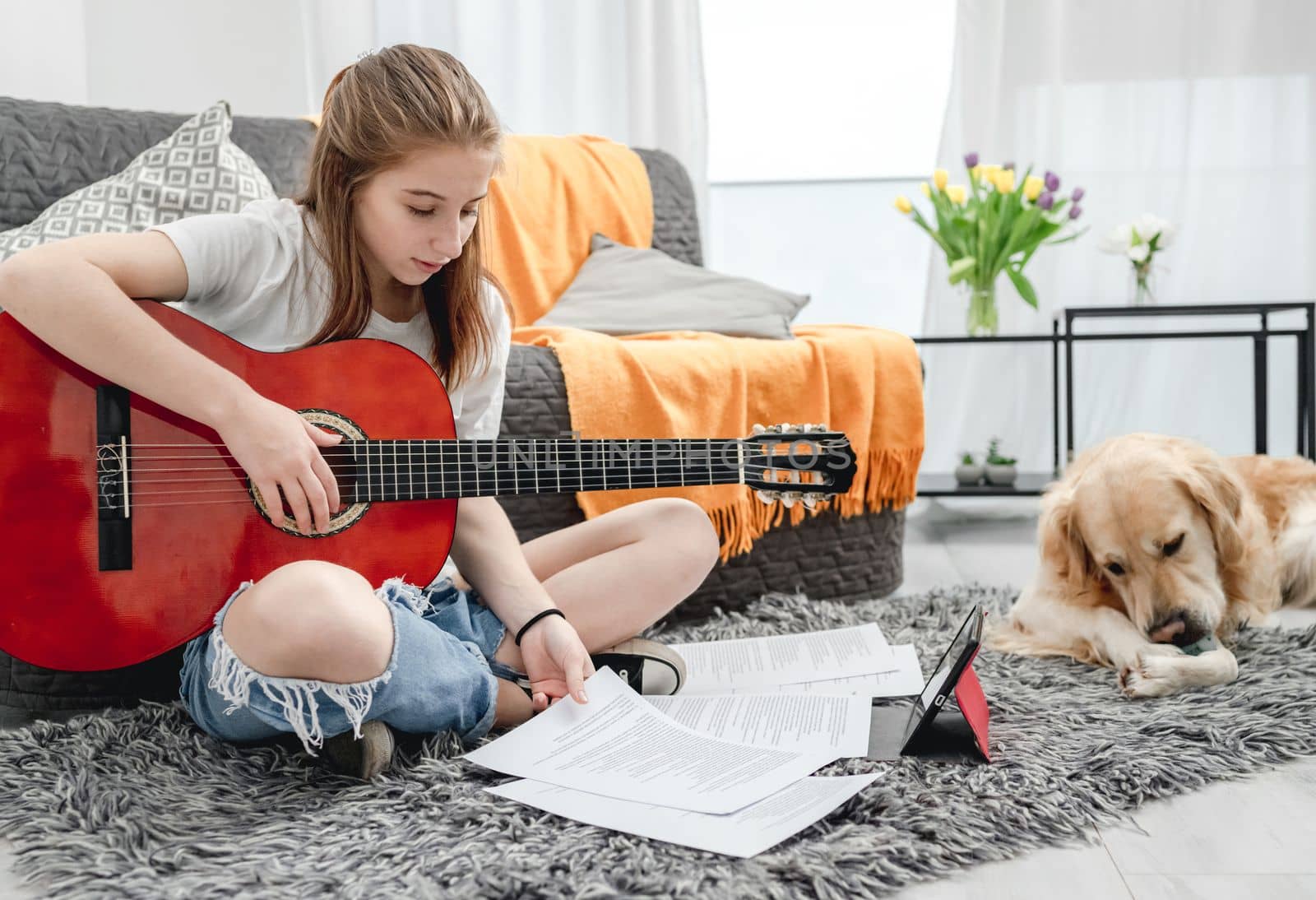 Girl teenager practicing guitar playing with golden retriever dog next to her at home sitting on floor. Pretty guitarist with musician instrument and purebred pet doggy