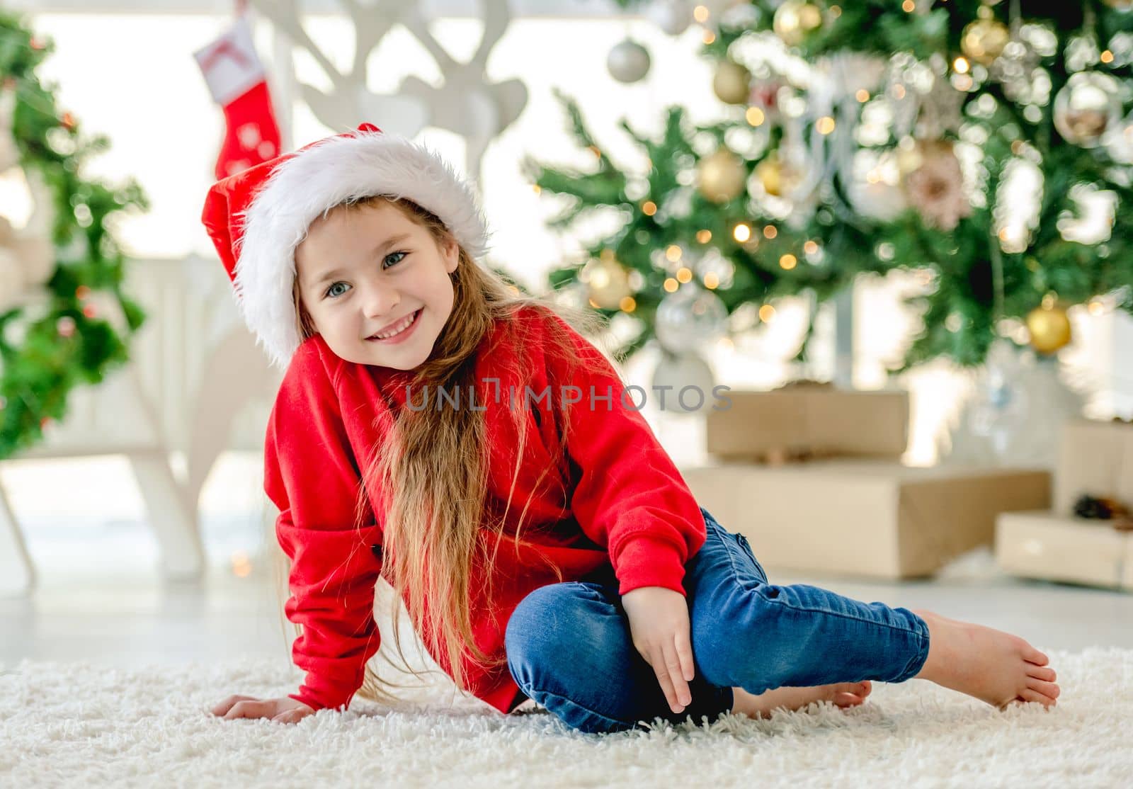 Little girl child wearing Santa hat in Christmas time posing on floor at decorated New Year home with festive tree. Cute kid in Xmas time