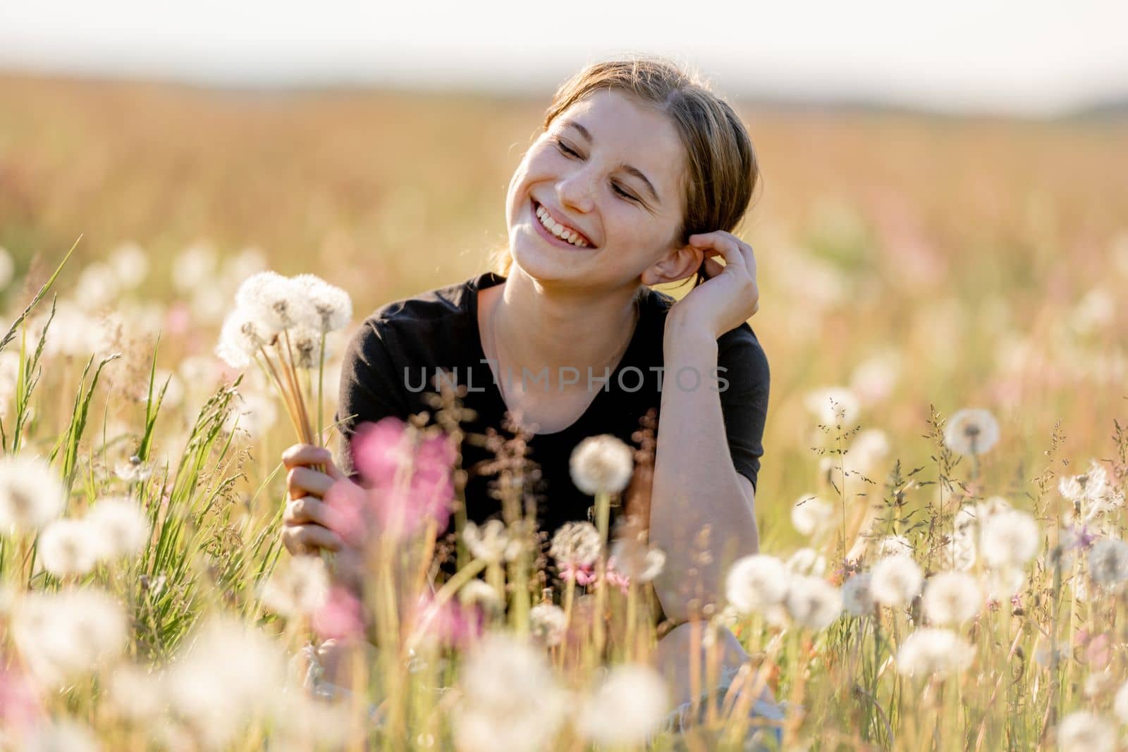 Pretty girl teenager sitting in field with flying dandelions and smiling. Beautiful young female person model at nature in sunny day portrait