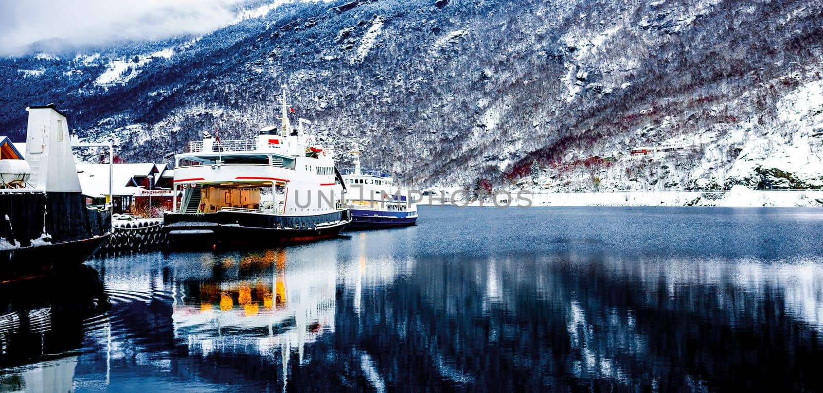 Cruise boat and traditional wooden houses in Norway surrounded with snowy mountains and cold lake in winer time. Beautiful norwegian nature, village and reflection in sea