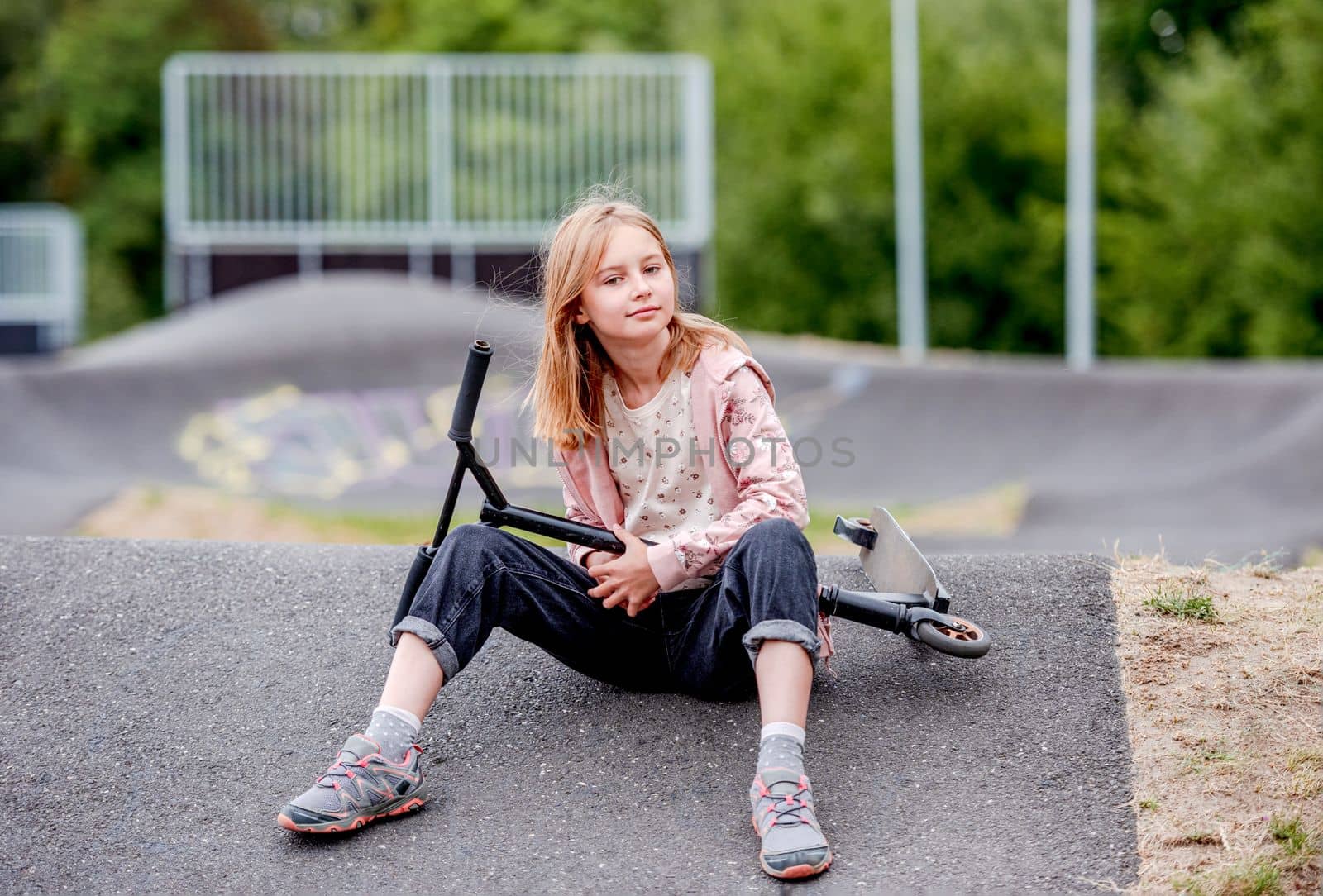 Preteen girl with scooter sitting in city park at spring time and looking at camera. Pretty child kid model posing with vehicle