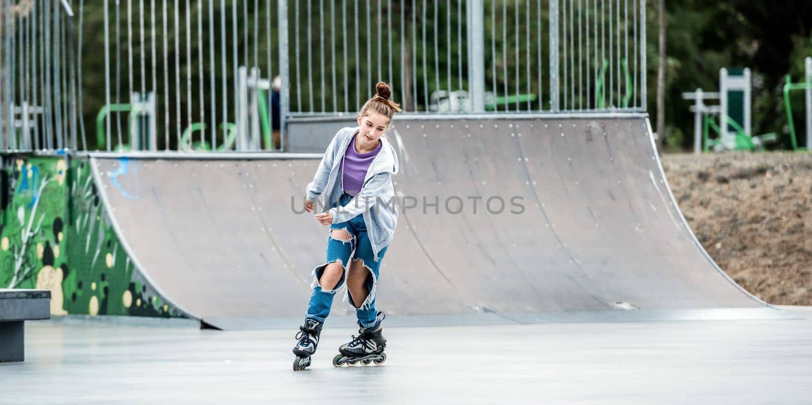 Beautiful girl roller skater riding in city park with ramp. Pretty female teenager rollerskating in casual clothes