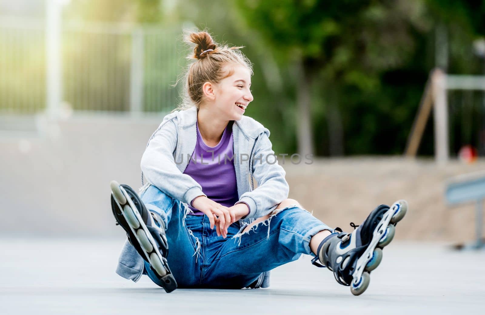 Cute girl roller skater sitting on ground in city park and smiling. Pretty female teenager with hairstyle posing during rollerskating