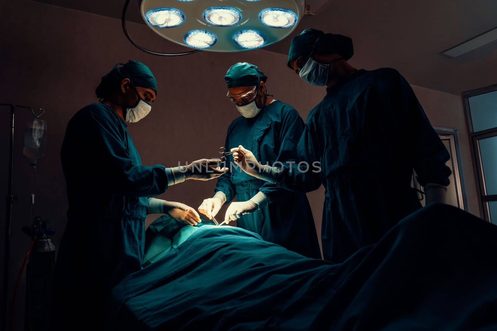 Surgical team performing surgery to patient in sterile operating room. In a surgery room lit by a lamp, a professional and confident surgical team provides medical care to an unconscious patient.