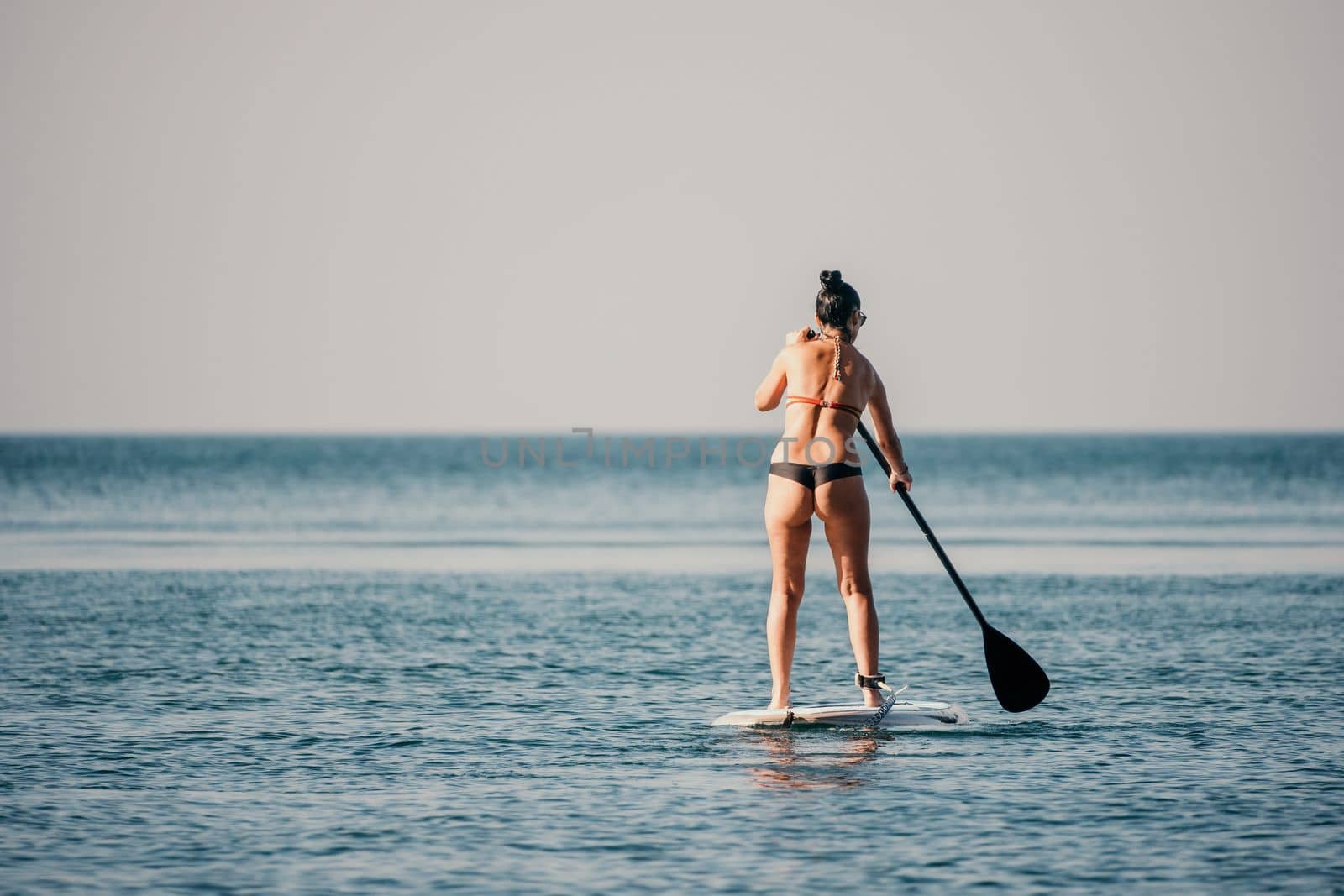 Sea woman sup. Silhouette of happy middle aged woman in rainbow bikini, surfing on SUP board, confident paddling through water surface. Idyllic sunset. Active lifestyle at sea or river
