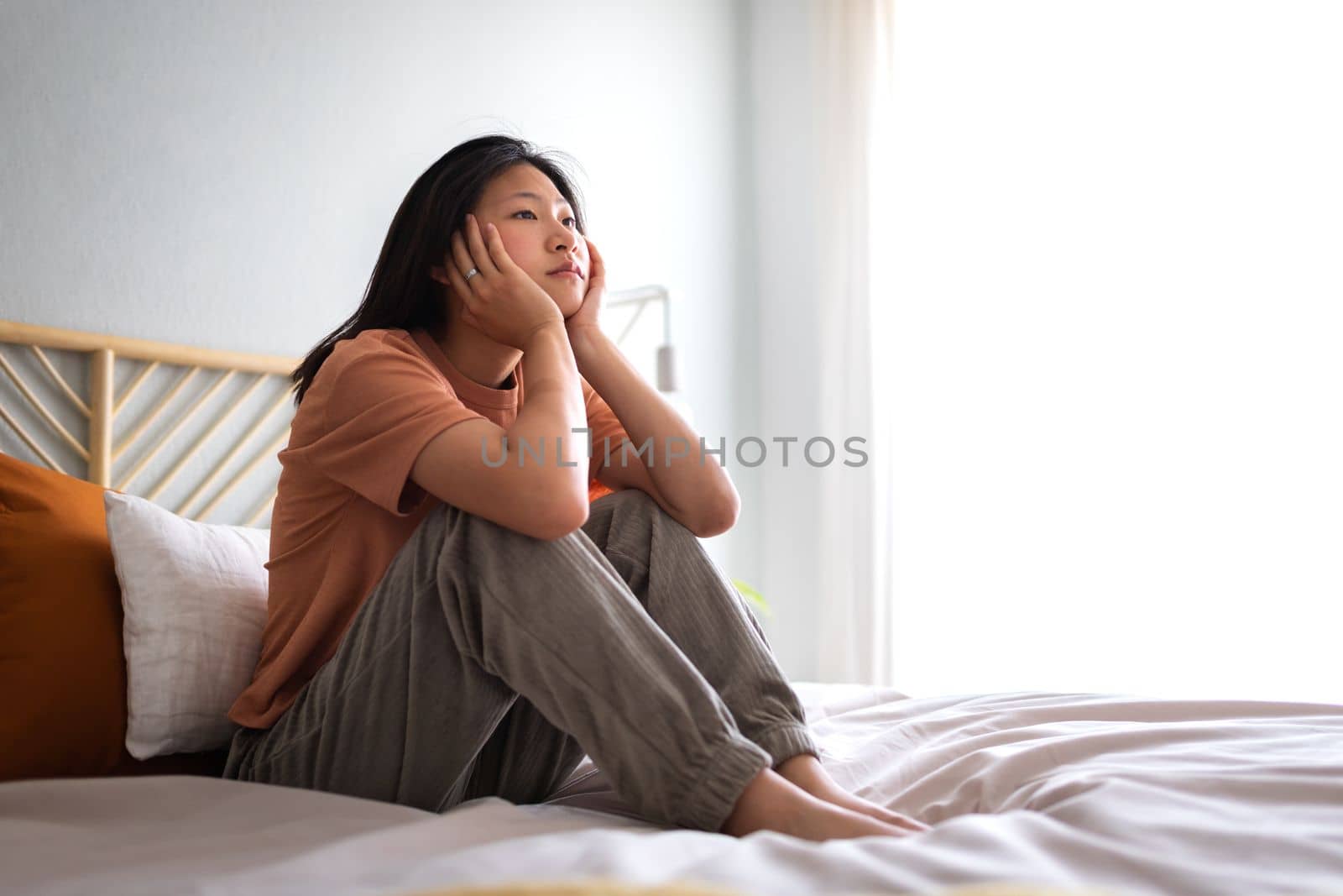 Pensive teen asian female sitting on bed. Bored young woman in cozy bedroom. Home loneliness. Copy space. Lifestyle concept.