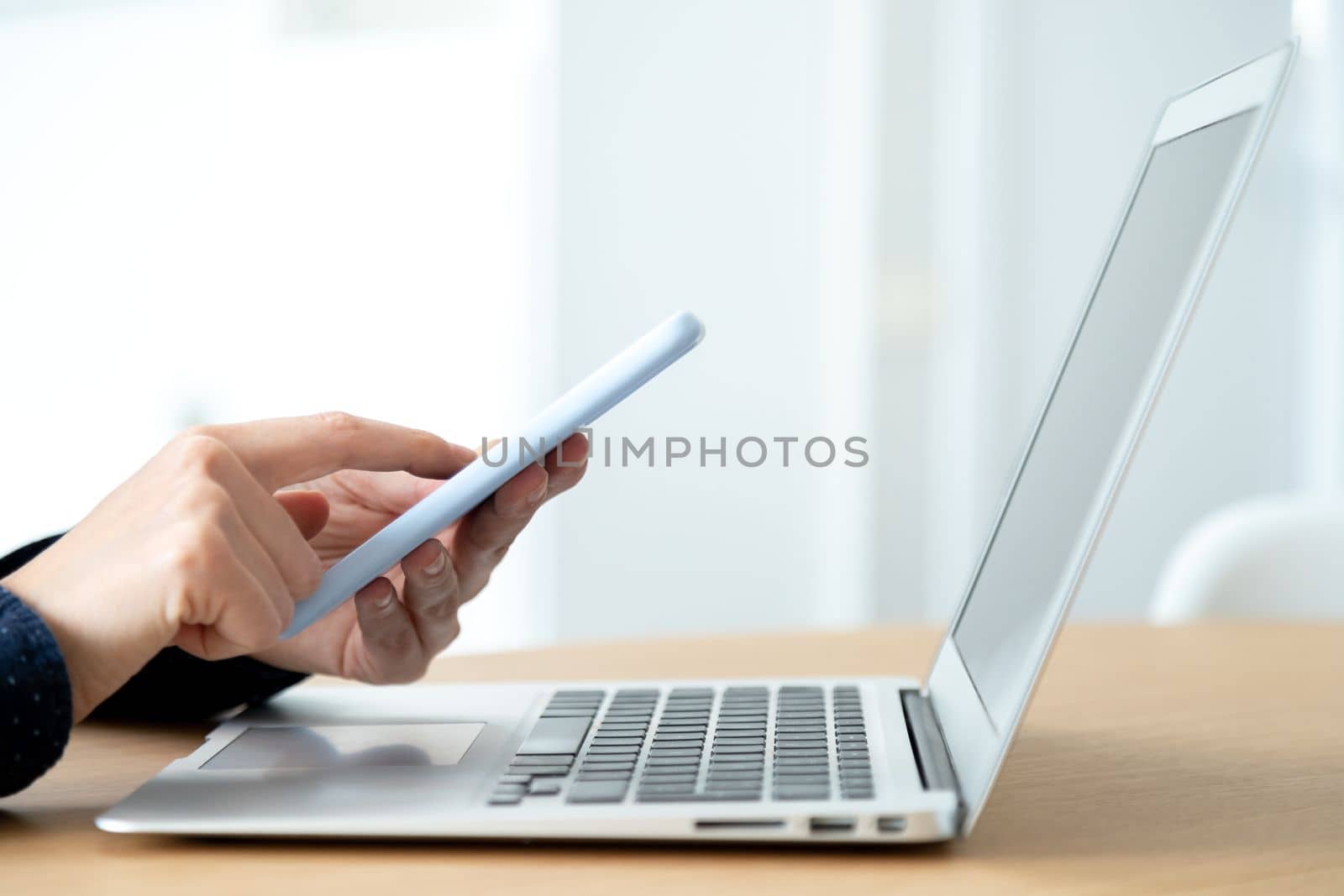 Side view of woman hands using mobile phone while working in the office with laptop. Blurred background. Texting concept.
