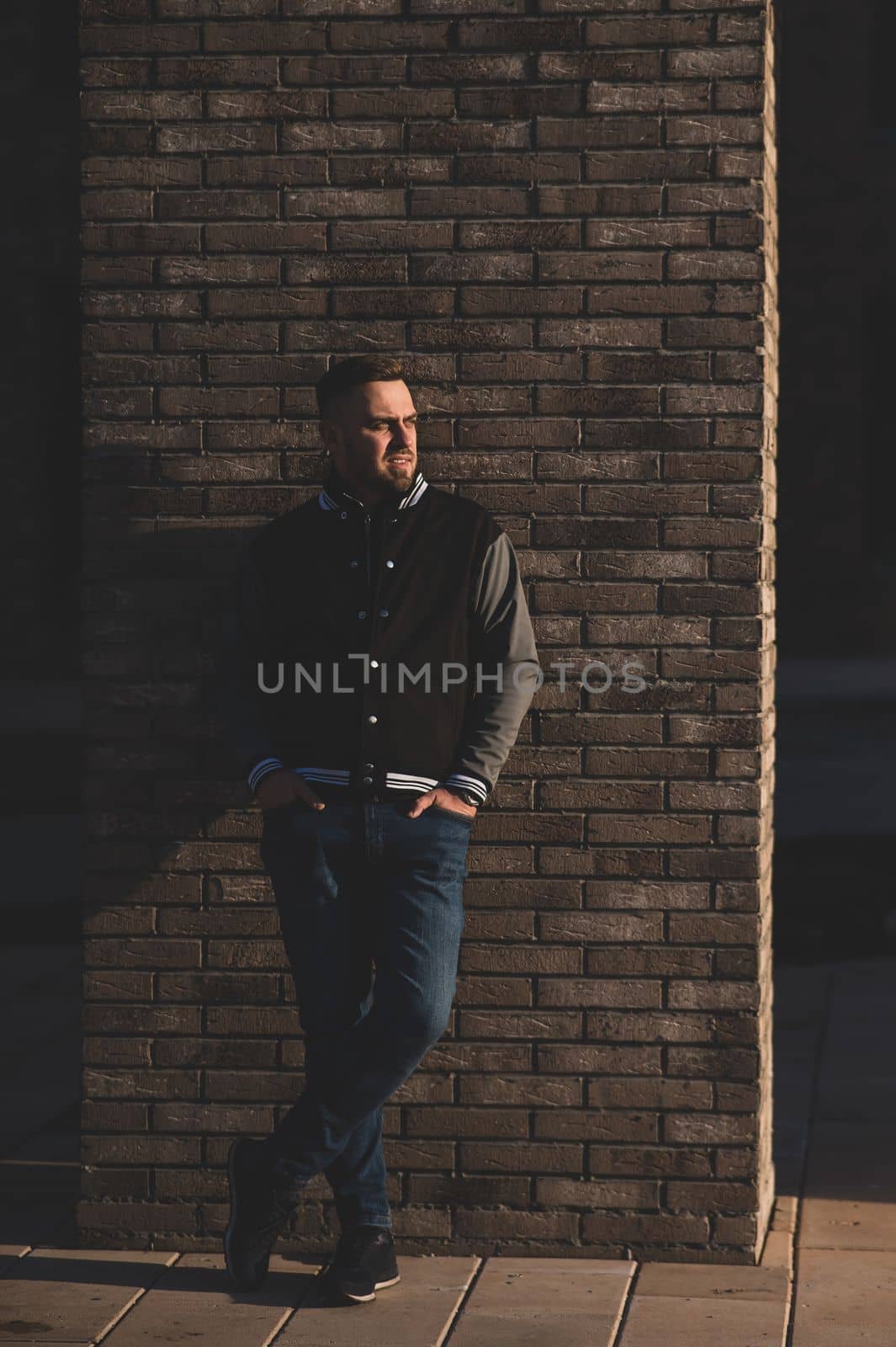 Caucasian bearded man in a bomber jacket leaned against a brick wall