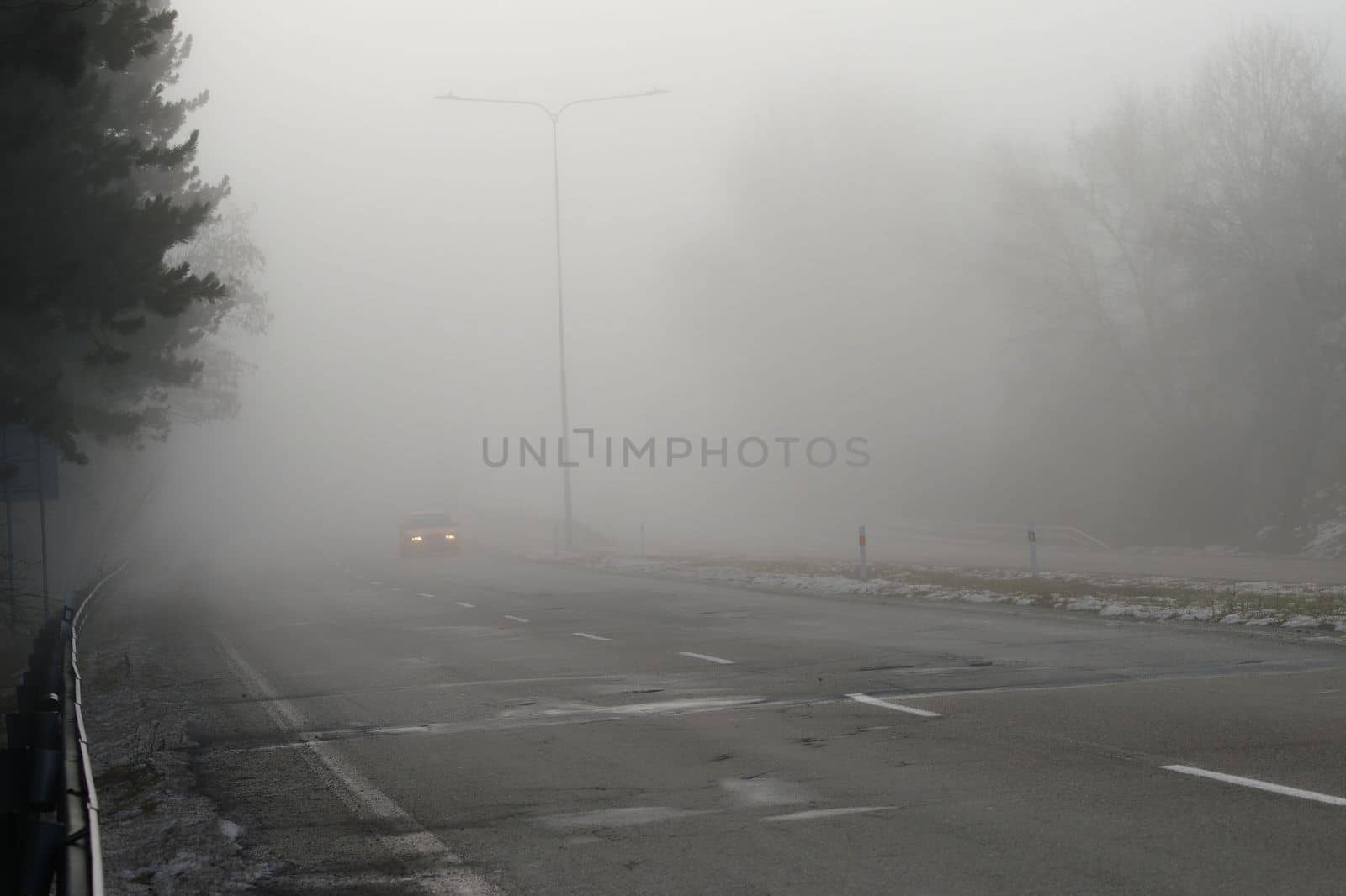 Car traveling on the foggy road with headlights or headlamps on. Low visibility - Dangerous driving of cars in winter in bad weather.