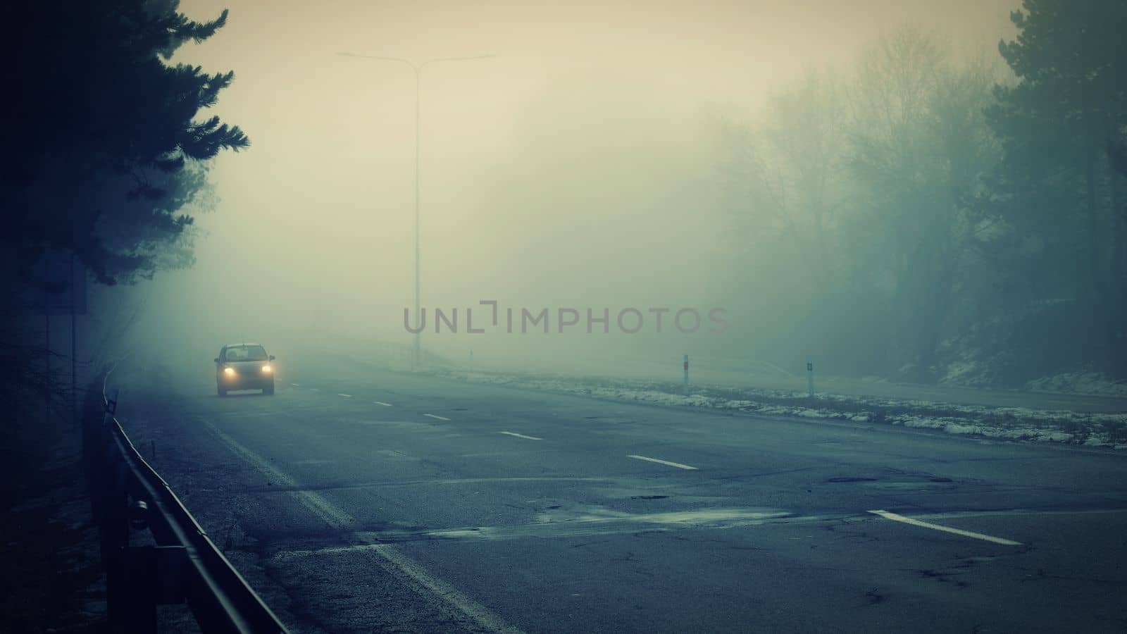 Car traveling on the foggy road with headlights or headlamps on. Low visibility - Dangerous driving of cars in winter in bad weather. by Montypeter