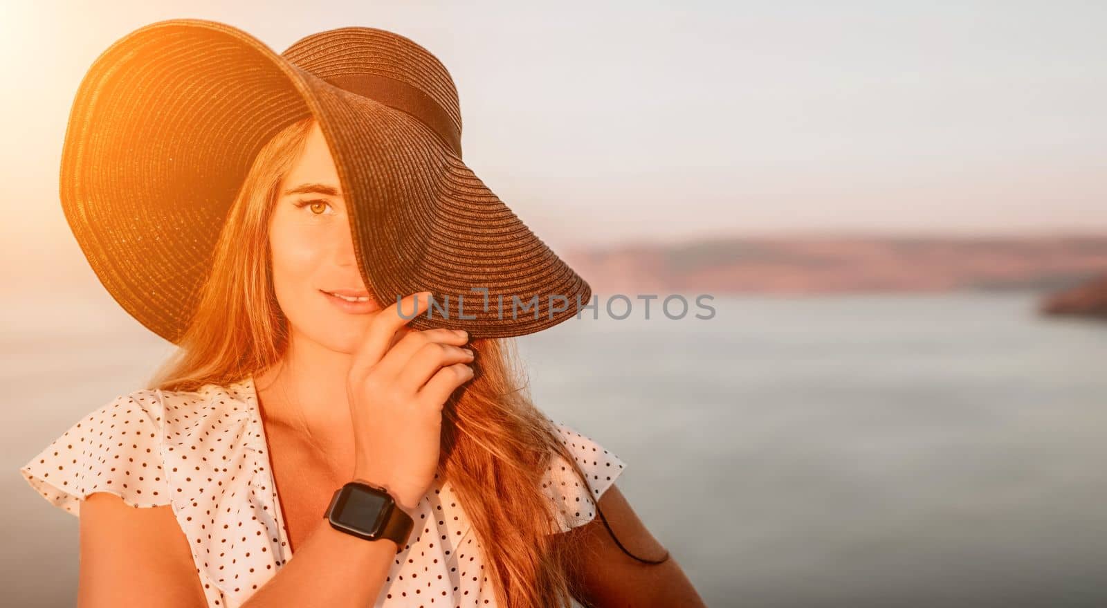 Portrait of happy young woman wearing summer black hat with large brim at beach on sunset. Sensual girl covering half face with hat on beach. Happy young woman smiling and looking at camera at sea. by panophotograph