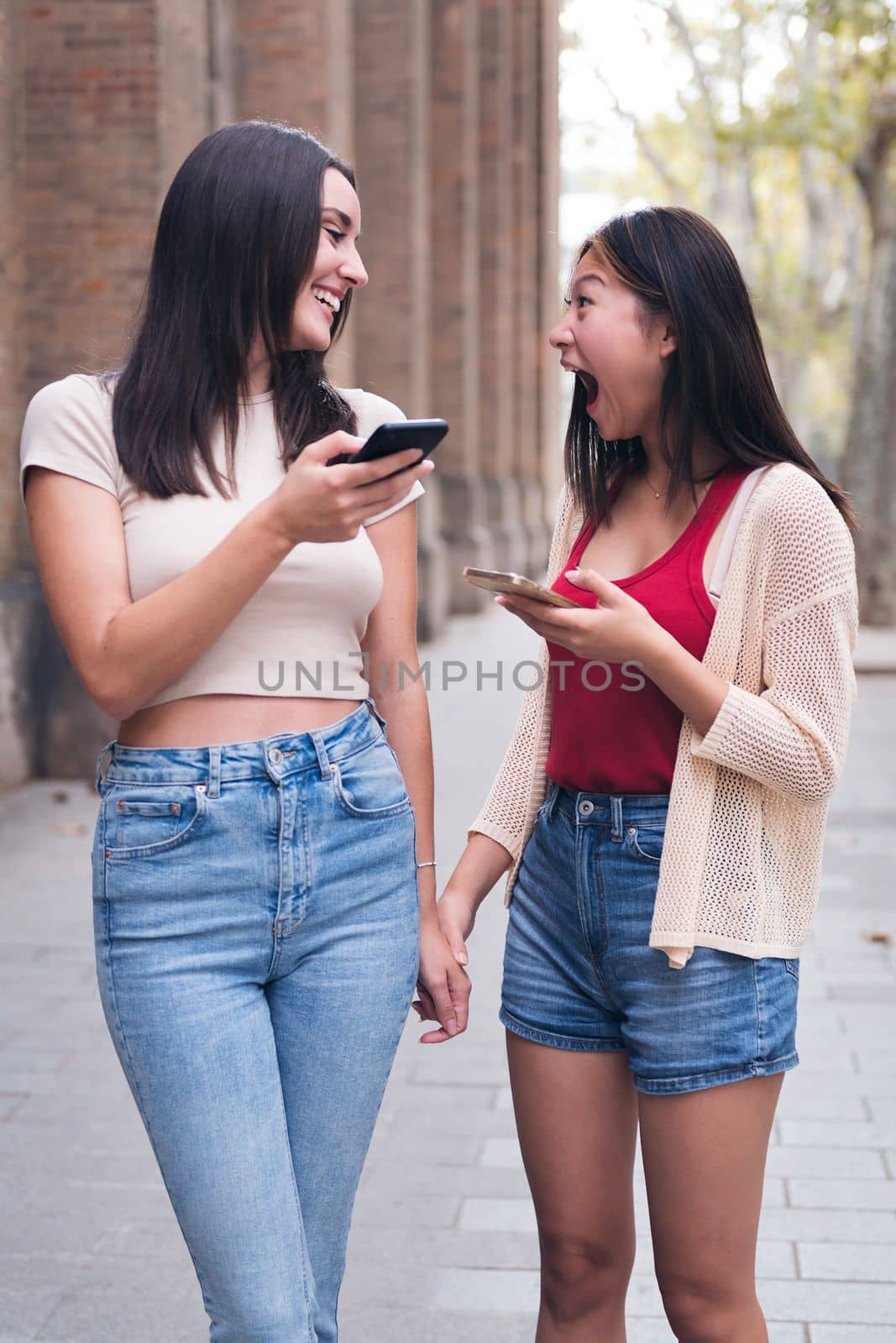 two young women sharing confidences and having fun with their cell phones while walking through the city holding hands, concept of friendship and love between people of the same sex