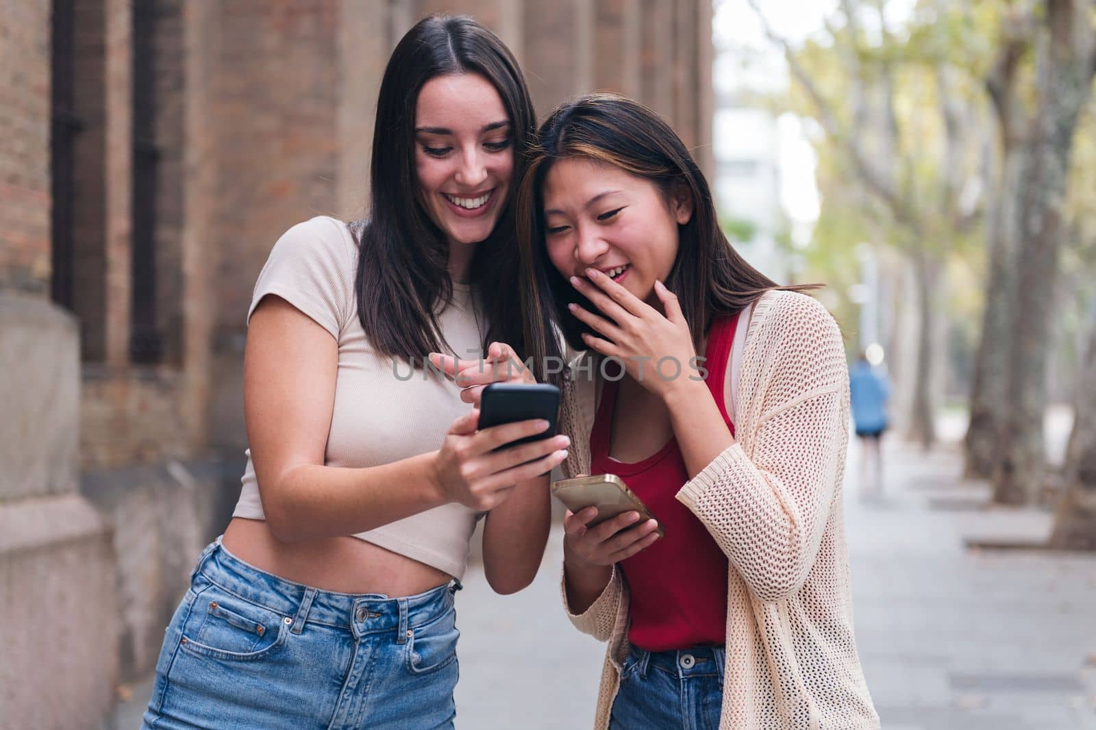 two young women sharing confidences and having fun with their cell phones while walking through the city, concept of friendship and love between people of the same sex