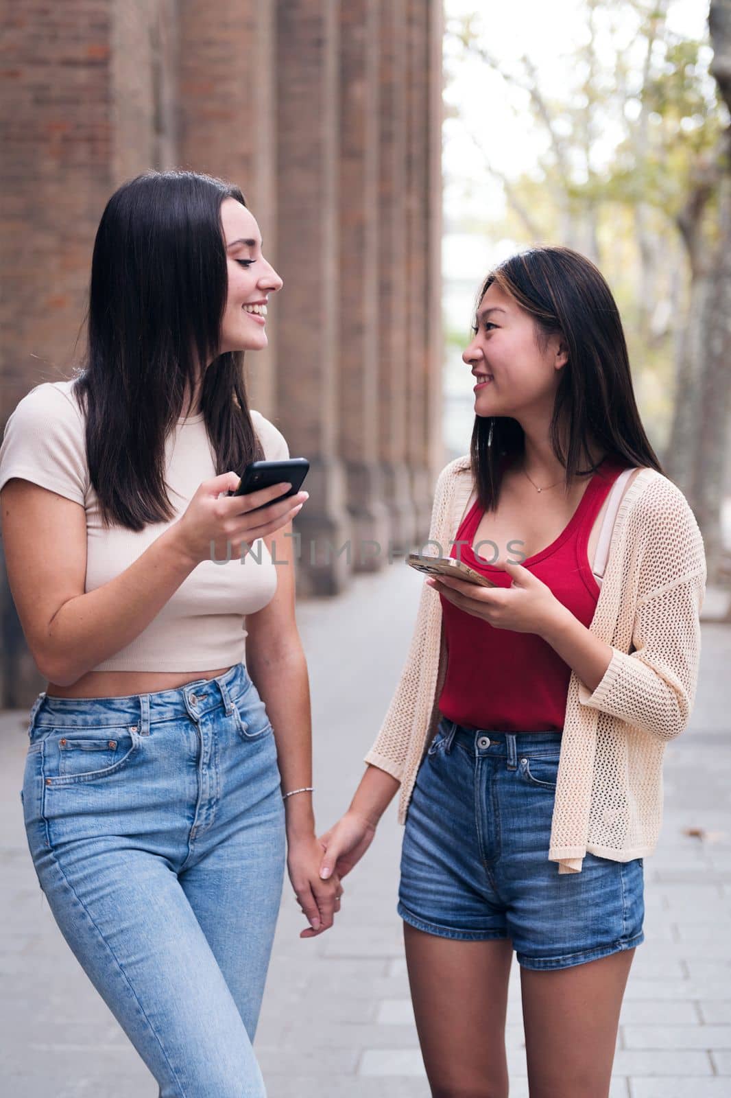 two young women having a good time with their cell phones while walking through the city holding hands, concept of friendship and love between people of the same sex