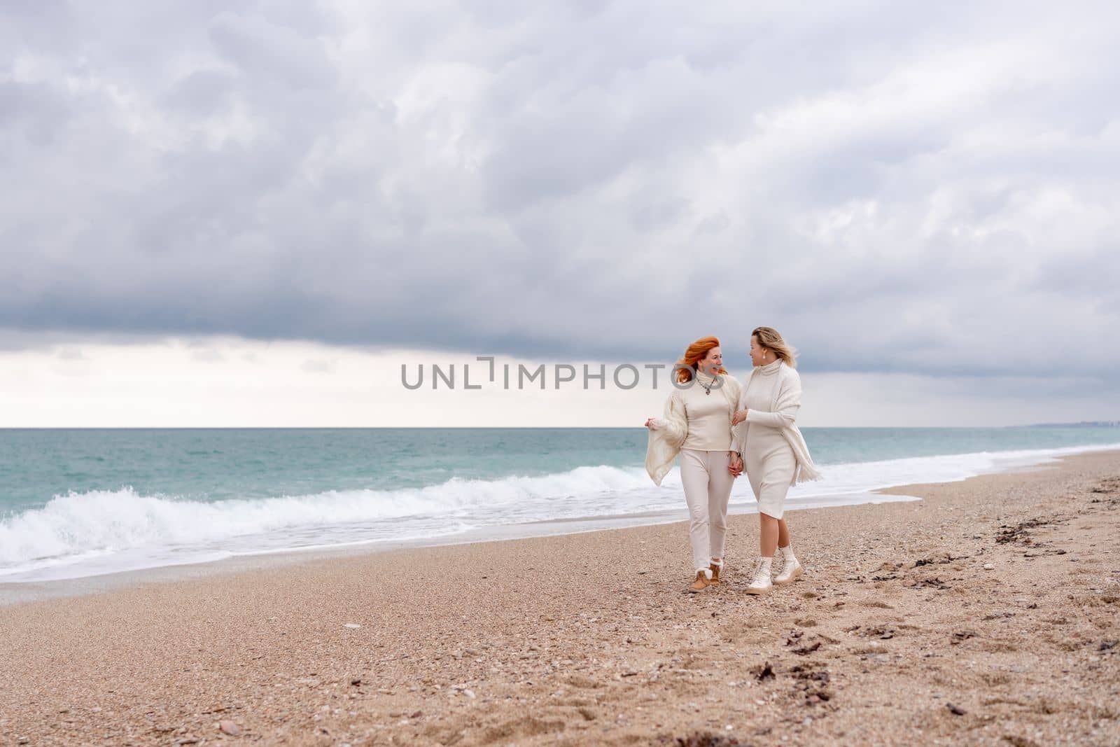 Women sea walk friendship spring. Two girlfriends, redhead and blonde, middle-aged walk along the sandy beach of the sea, dressed in white clothes. Against the backdrop of a cloudy sky and the winter sea. Weekend concept. by Matiunina