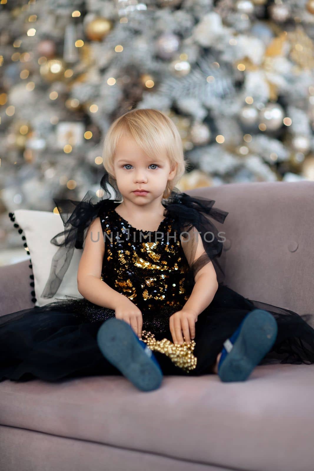 A little girl is sitting on the background of a Christmas tree. Blonde 2 years old in a black dress sits on a beige sofa in a room decorated for christmas and new year. Photo under the tree