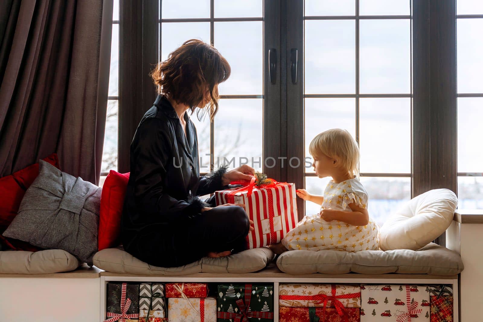 Mother and daughter sit on the windowsill and hold a gift between them. In a decorated Christmas room, mother in black pajamas, daughter in white dress. New Year's family holidays. by Matiunina