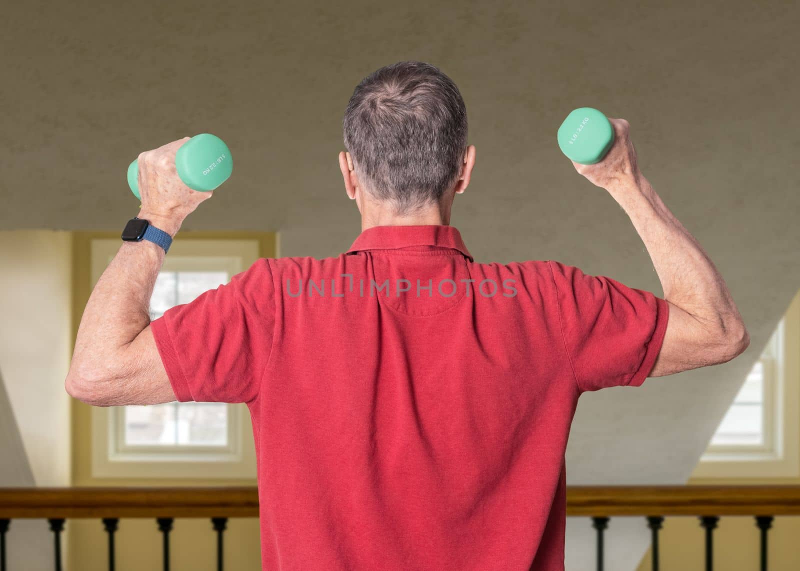 Senior man in red shirt seen from back and exercising with dumbbells in home against railing