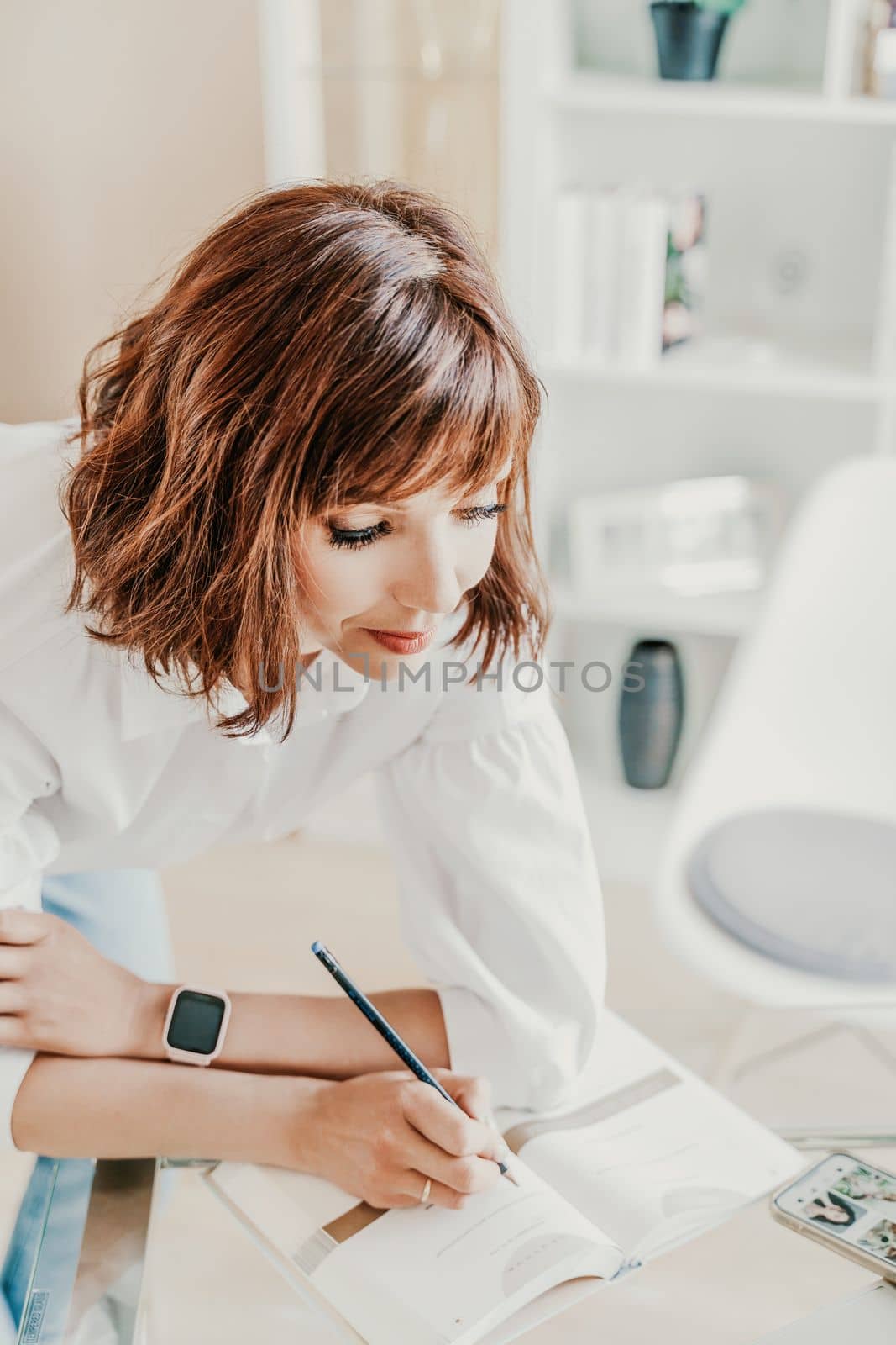 Business woman in bright office. A woman writes in a notebook while standing near the workplace desk. A business woman working at a desk in an office is dressed in jeans and a white shirt. by Matiunina