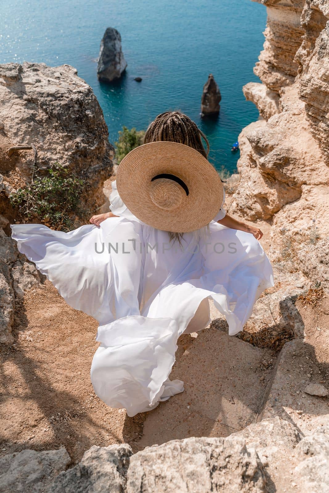 Woman white dress sea rocks. A girl in a hat and a long white dress descends the stairs between the yellow rocks overlooking the sea. The stone can be seen in the sea. Travel photography. by Matiunina
