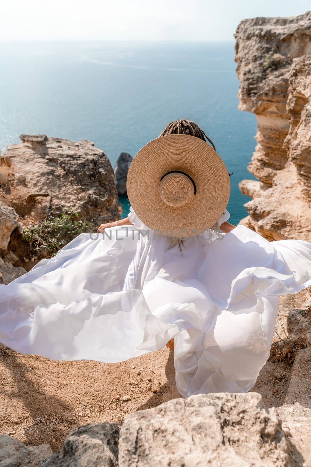 Woman white dress sea rocks. A girl in a hat and a long white dress descends the stairs between the yellow rocks overlooking the sea. The stone can be seen in the sea. Travel photography. by Matiunina