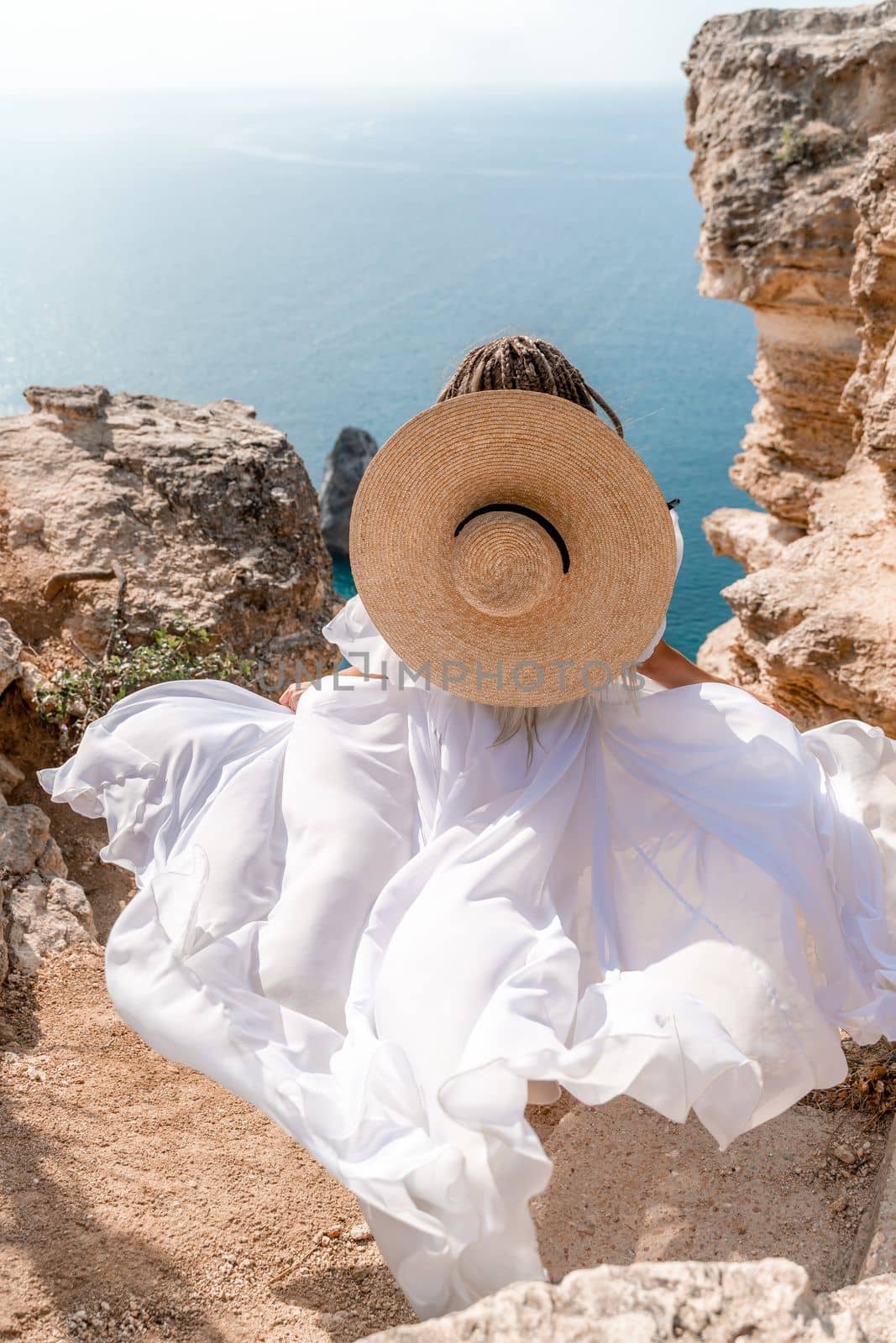 Woman white dress sea rocks. A girl in a hat and a long white dress descends the stairs between the yellow rocks overlooking the sea. The stone can be seen in the sea. Travel photograph