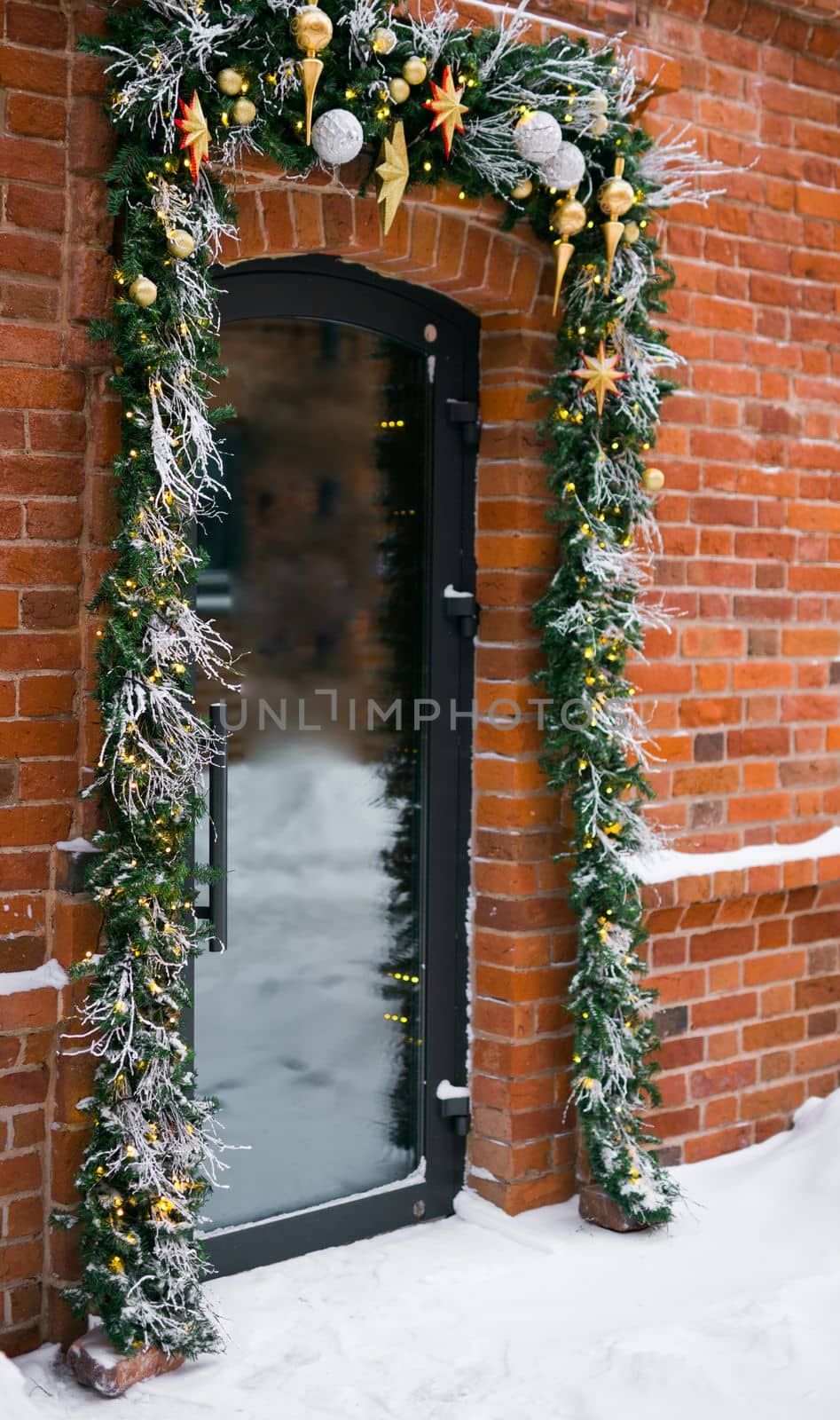 Stylish christmas fir branches with red baubles and sparkling garland on front of door at holiday market or restaurant in city street. Winter christmas street decor