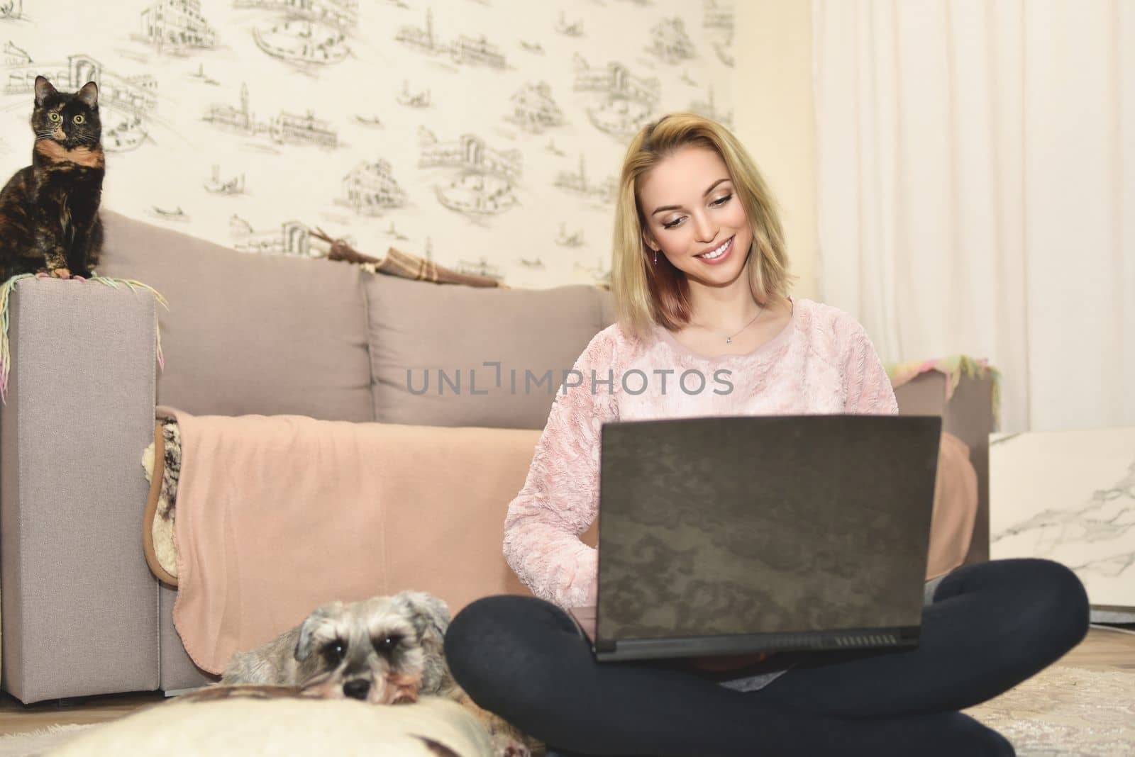 woman smiles during a video call on a laptop, little cat sitting on the couch and cute dog is sleeping on a pillow nearby.