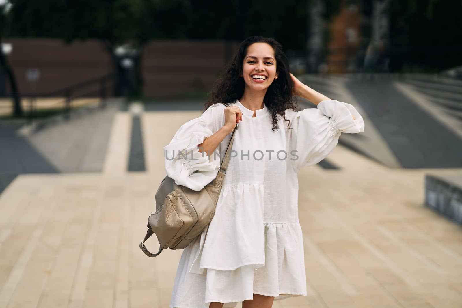 Smiling curly brunette girl in a white dress with a backpack putting her hand behind her head by driver-s